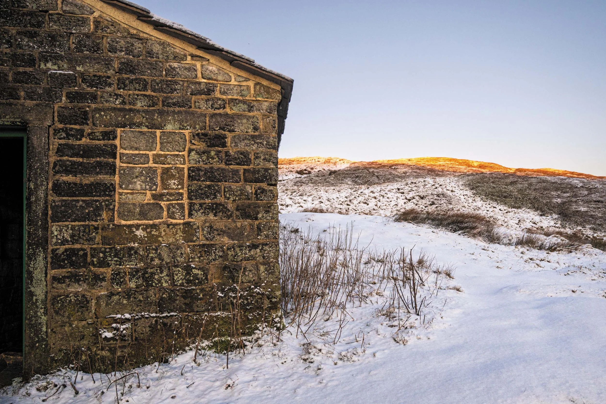 Een stenen muur van een oud gebouw met sneeuw op de grond en een berglandschap op de achtergrond tijdens zonsondergang.