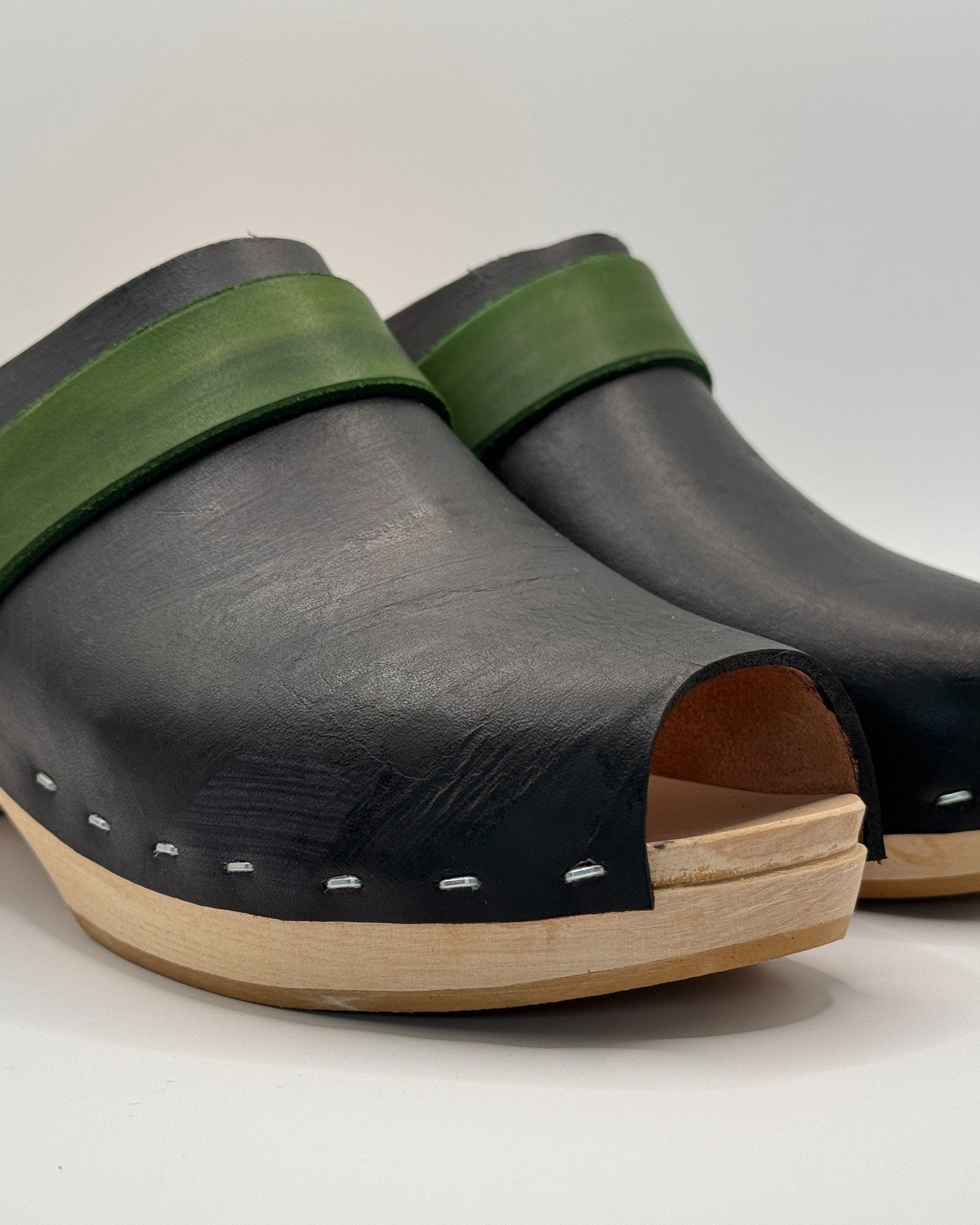 These handmade black peep-toe clogs with a green strap offer a unique fusion of classic elegance and bold contrast. The sleek black leather upper provides a timeless and versatile look, while the vibrant green strap adds a playful yet sophisticated t