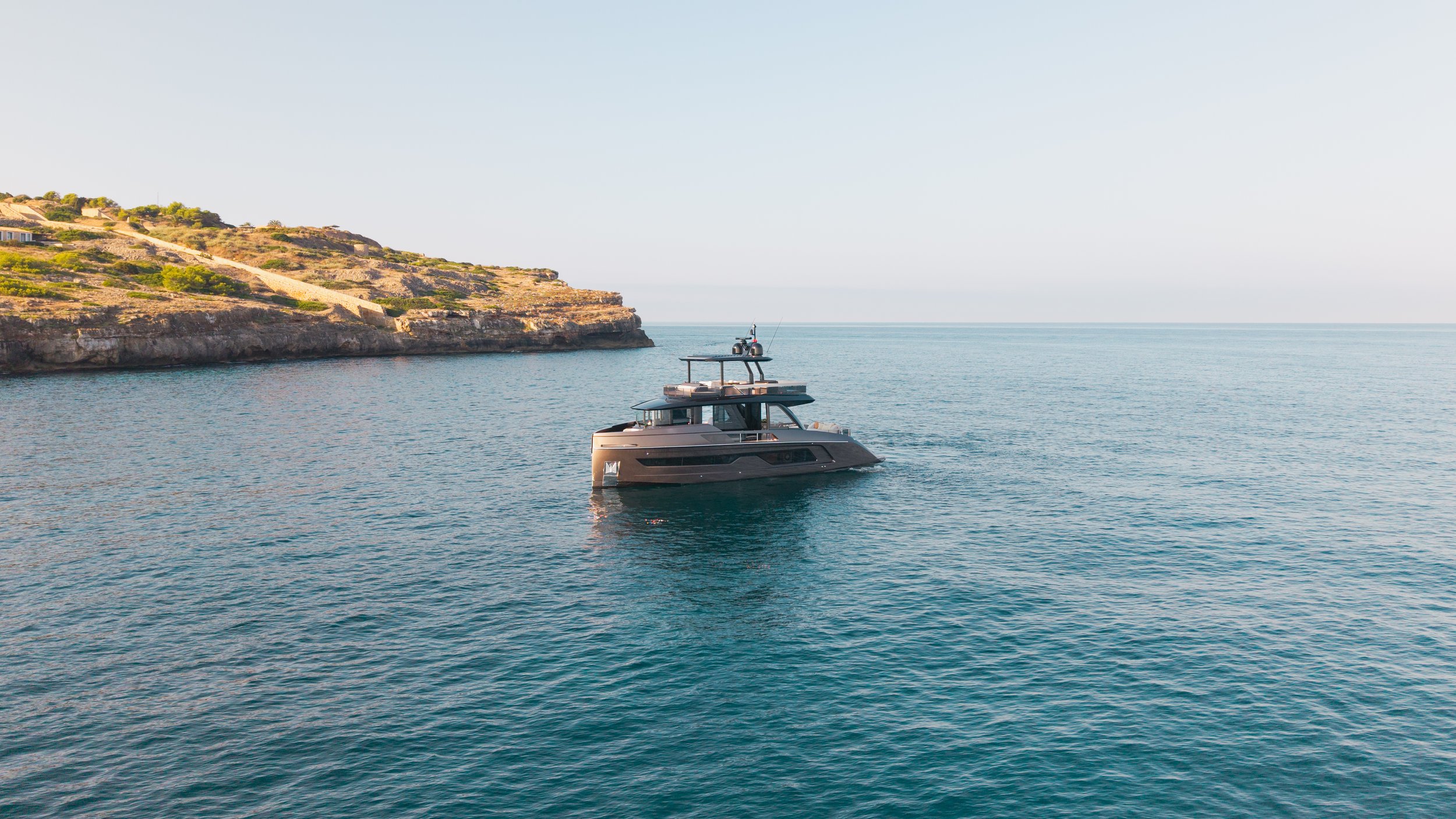 A luxury yacht floating on calm ocean waters near a rocky coastline with some houses on the hill in the background.