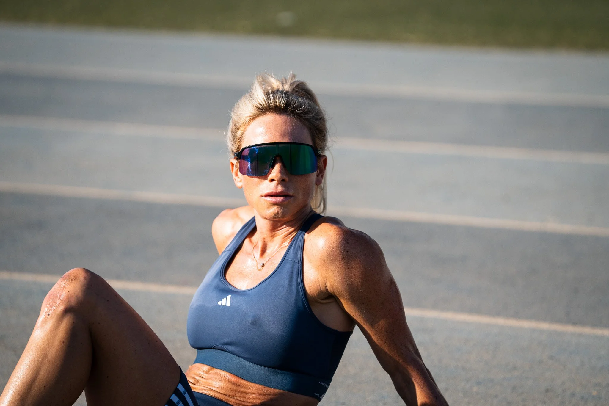 Female athlete wearing sunglasses and a blue sports top, sitting on the ground on a running track.