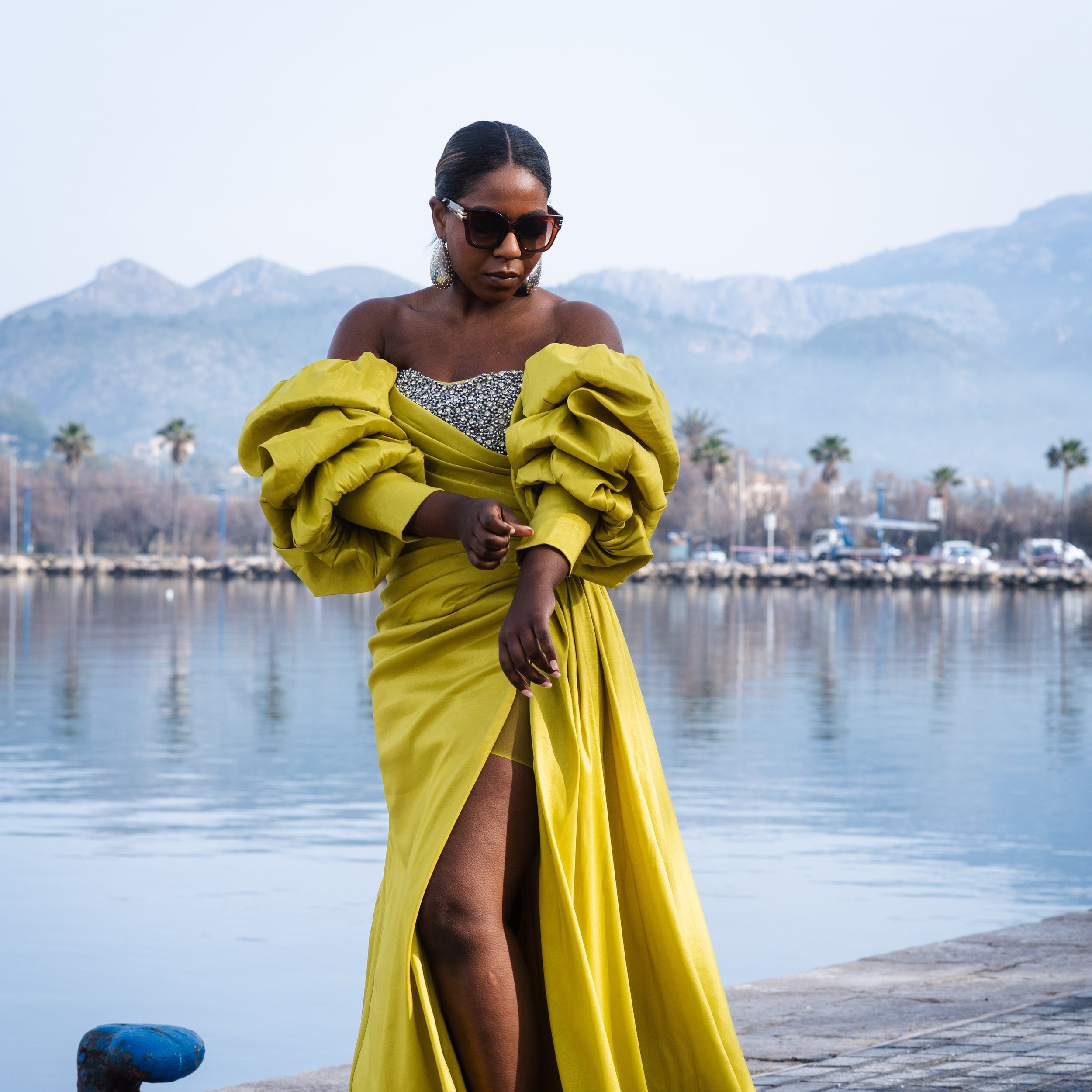 Person wearing a yellow dress with dramatic sleeves and sunglasses standing near a waterfront, with mountains and palm trees in the background.