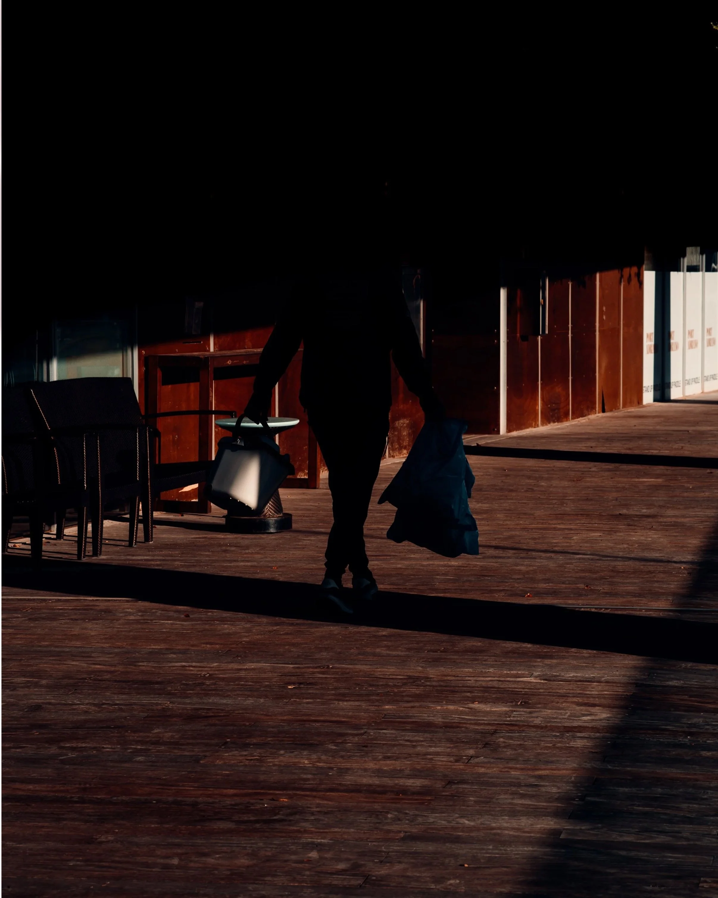 Silhouette of a person carrying bags along a shaded wooden walkway.
