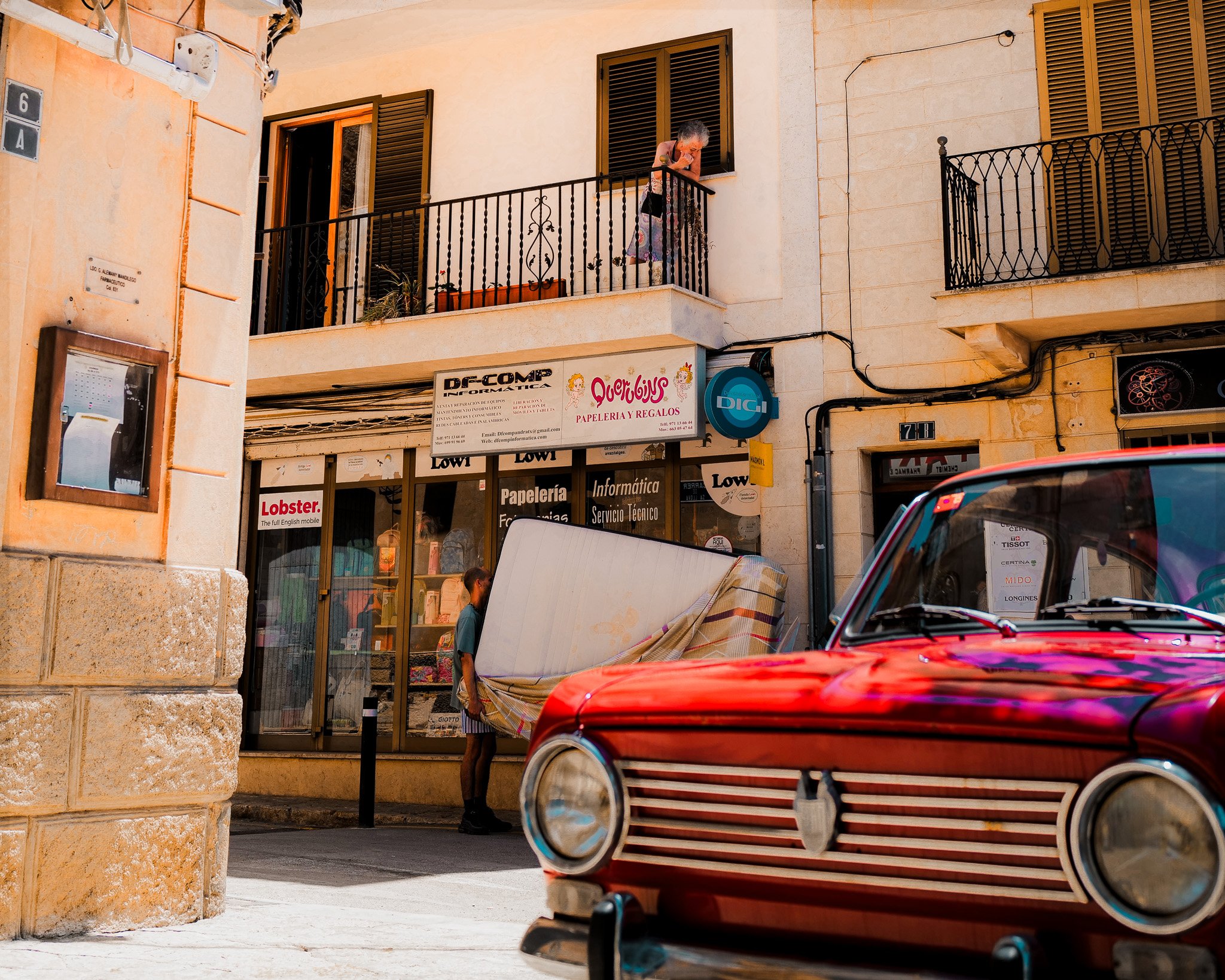 Street scene with a red vintage car parked in front of shops. A person is carrying a large item wrapped in a striped cloth. A person on a balcony is looking down at the street. Store signs are visible, including "Lobster" and "DF-COMP."