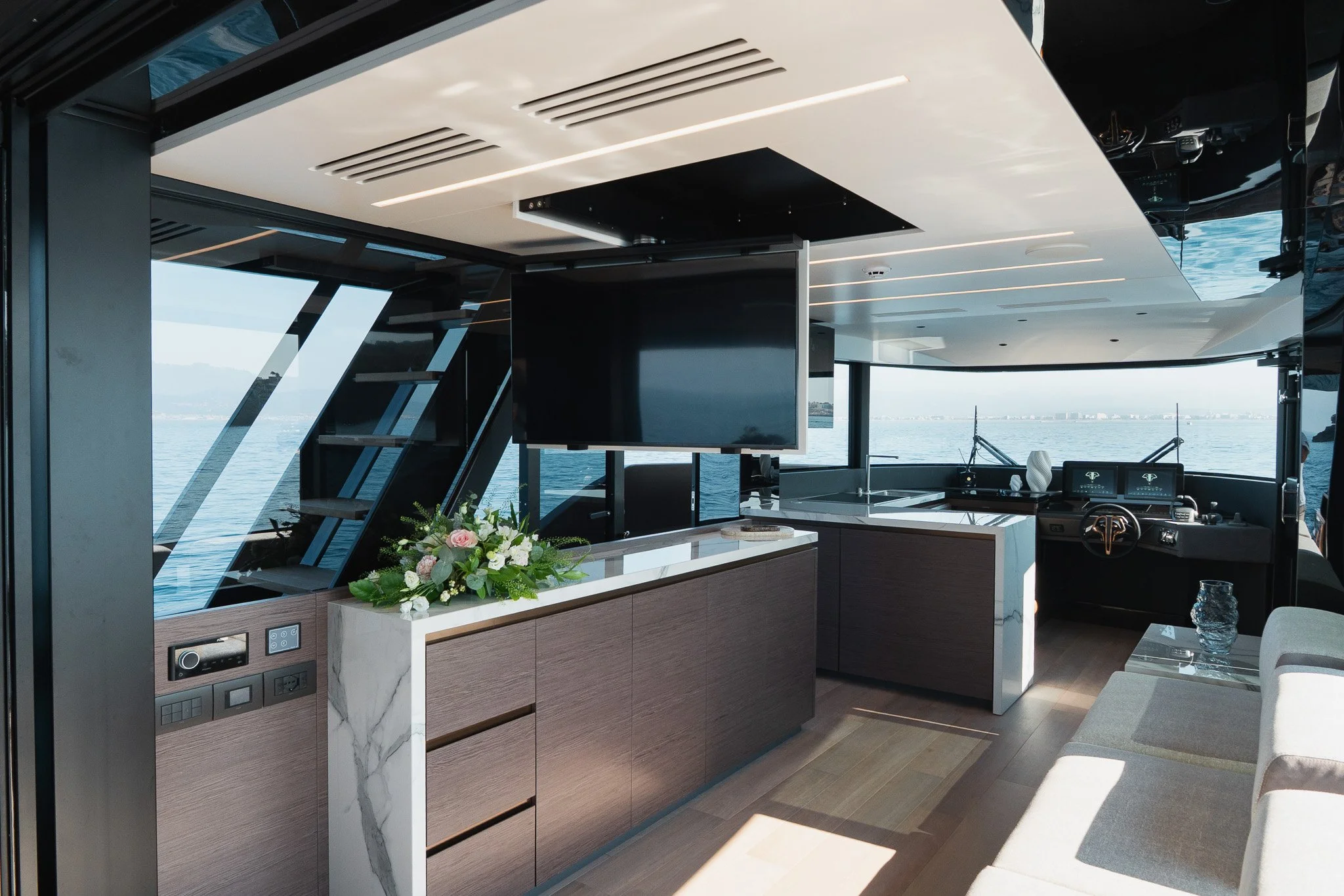 Interior of a luxury yacht with a modern design, featuring a flat-screen TV, a floral arrangement, a steering wheel, and large windows overlooking water.