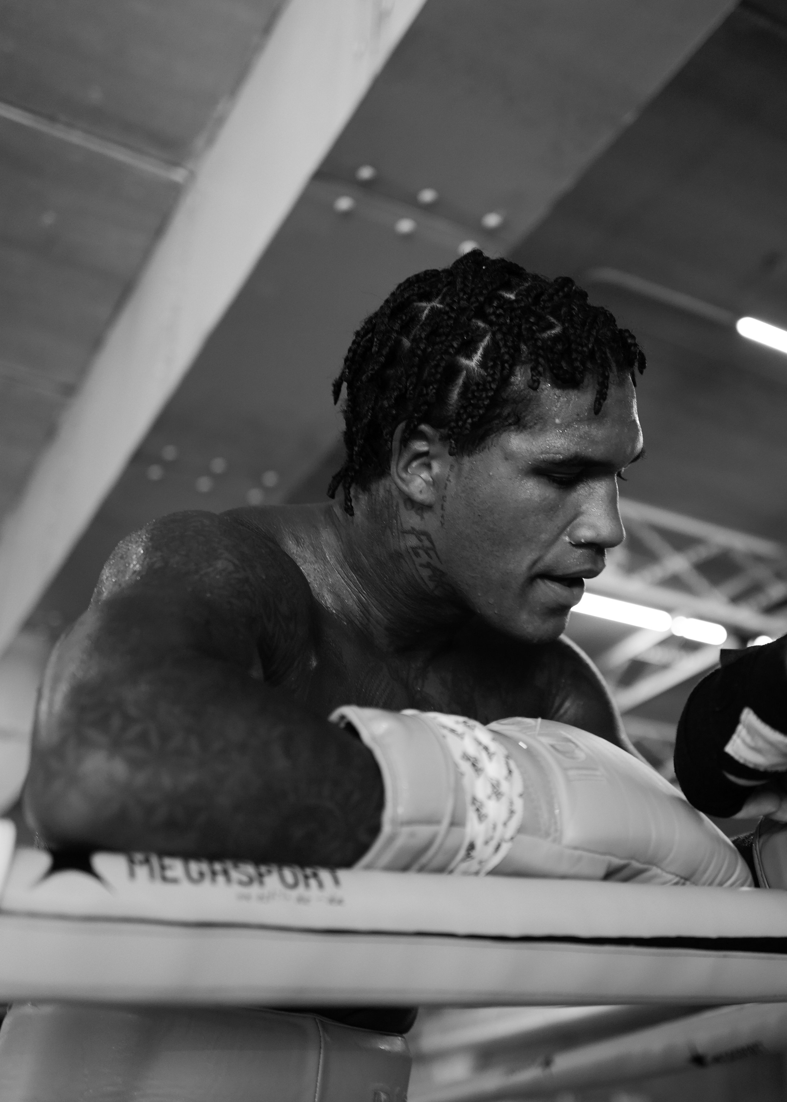 A male boxer with braided hair, tattoos, and a focused expression resting on a boxing ring's corner pad, wearing boxing gloves.