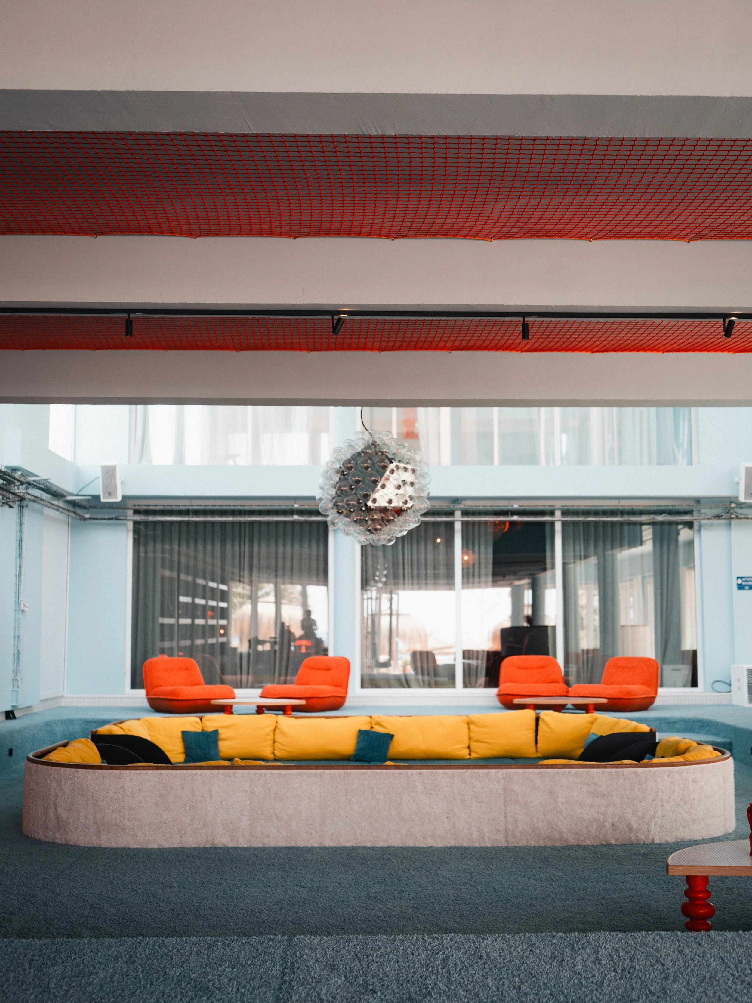 Modern interior lounge area with yellow curved sofa, orange chairs, and contemporary chandelier, with glass windows and blue walls.