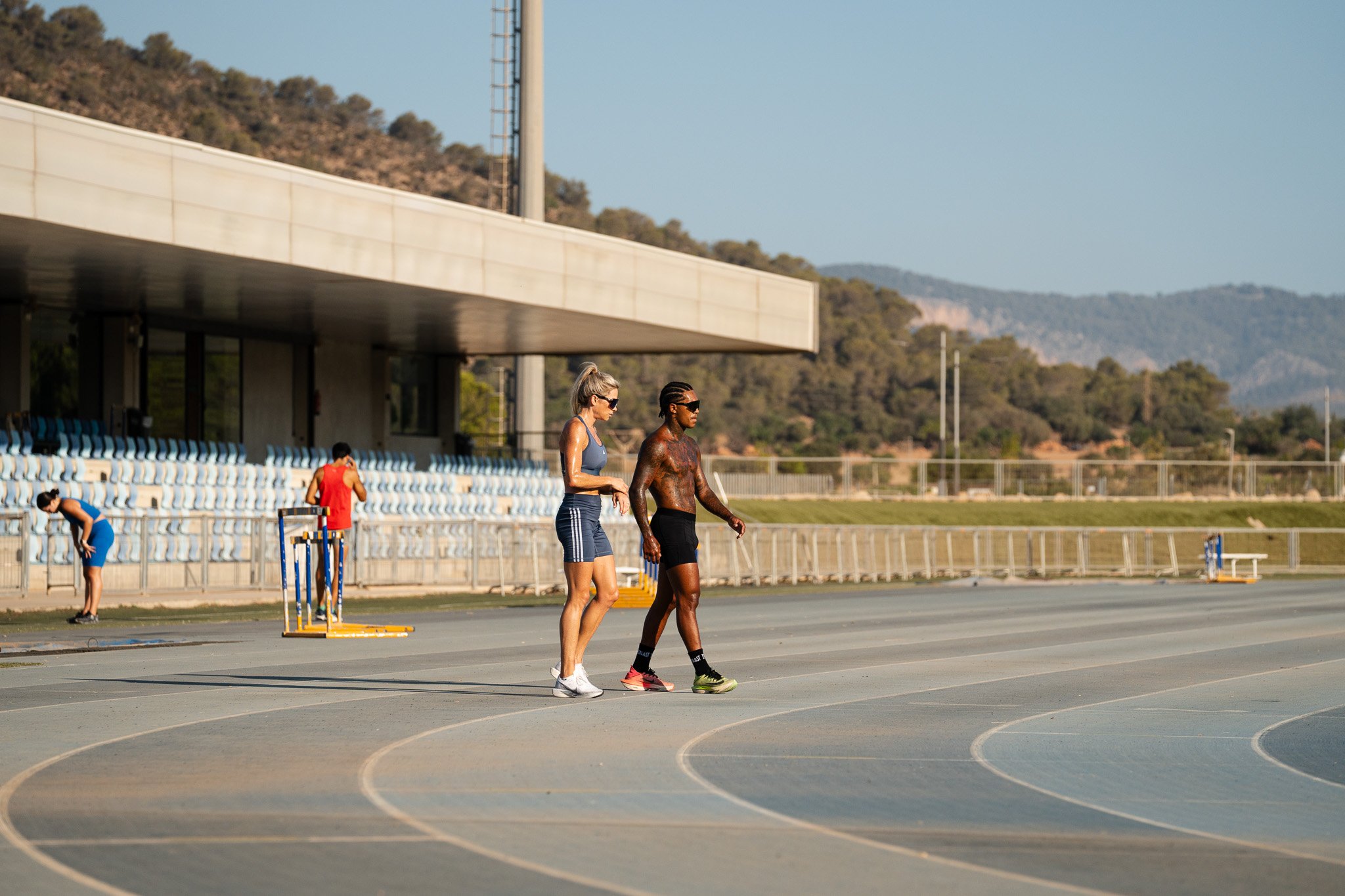 Two people walking on an outdoor running track near bleachers, with a forested hill in the background. A hurdle and two other people are in the distance, one of whom is wearing blue and bending forward.