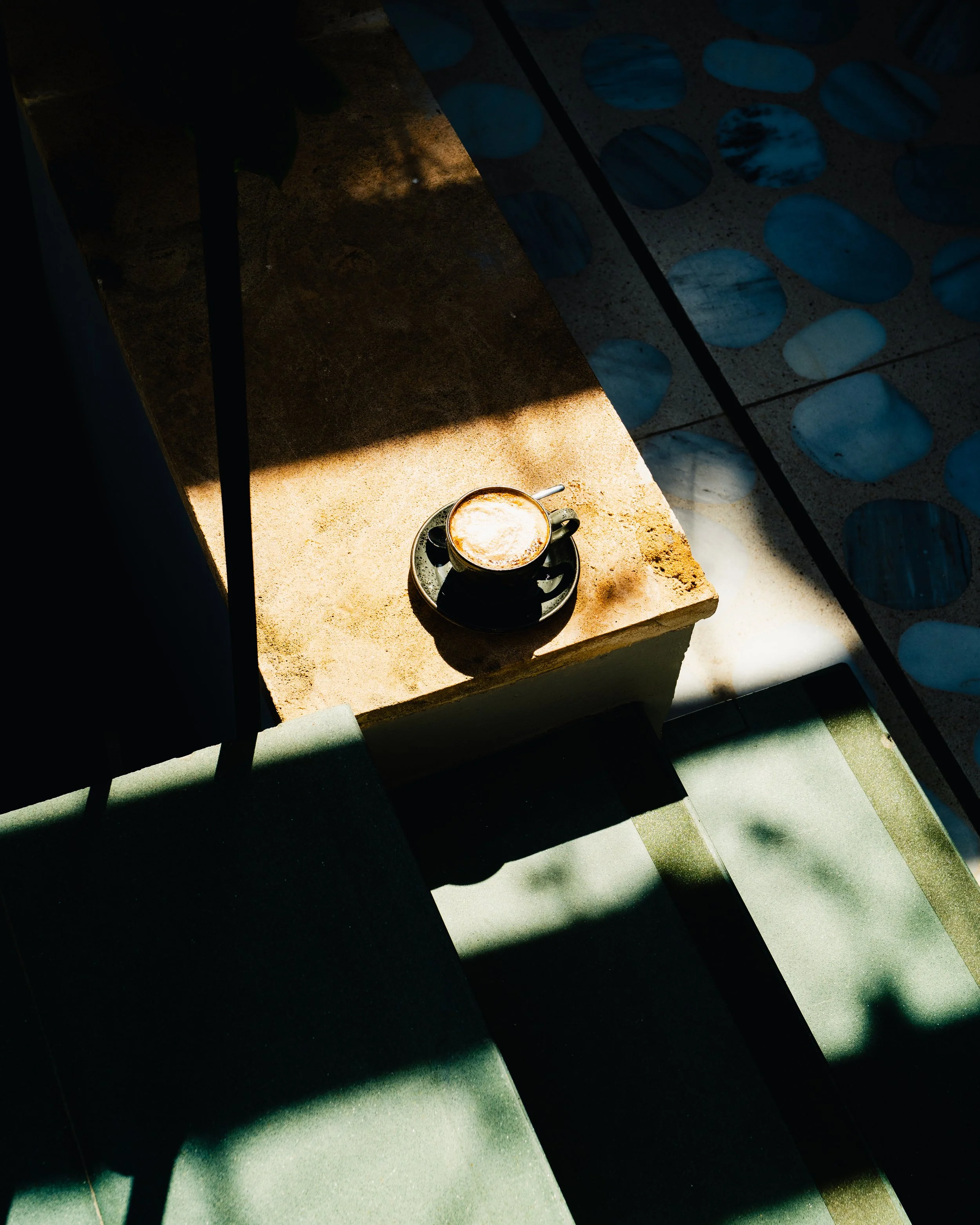 A black coffee cup on a saucer with a spoon, sitting on a beige concrete ledge. Natural sunlight creates shadows across the ledge and surrounding surfaces, with patterned blue and white tiles in the background.