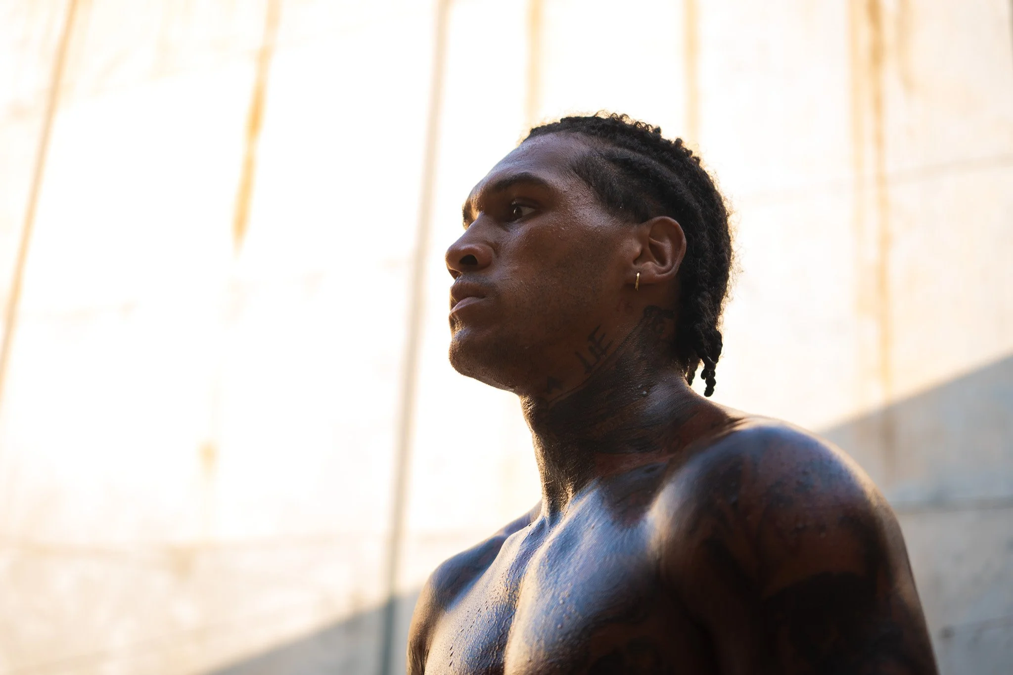 Profile of a shirtless man with tattoos, earrings, and dreadlocks, with a concrete wall background, captured in natural lighting.