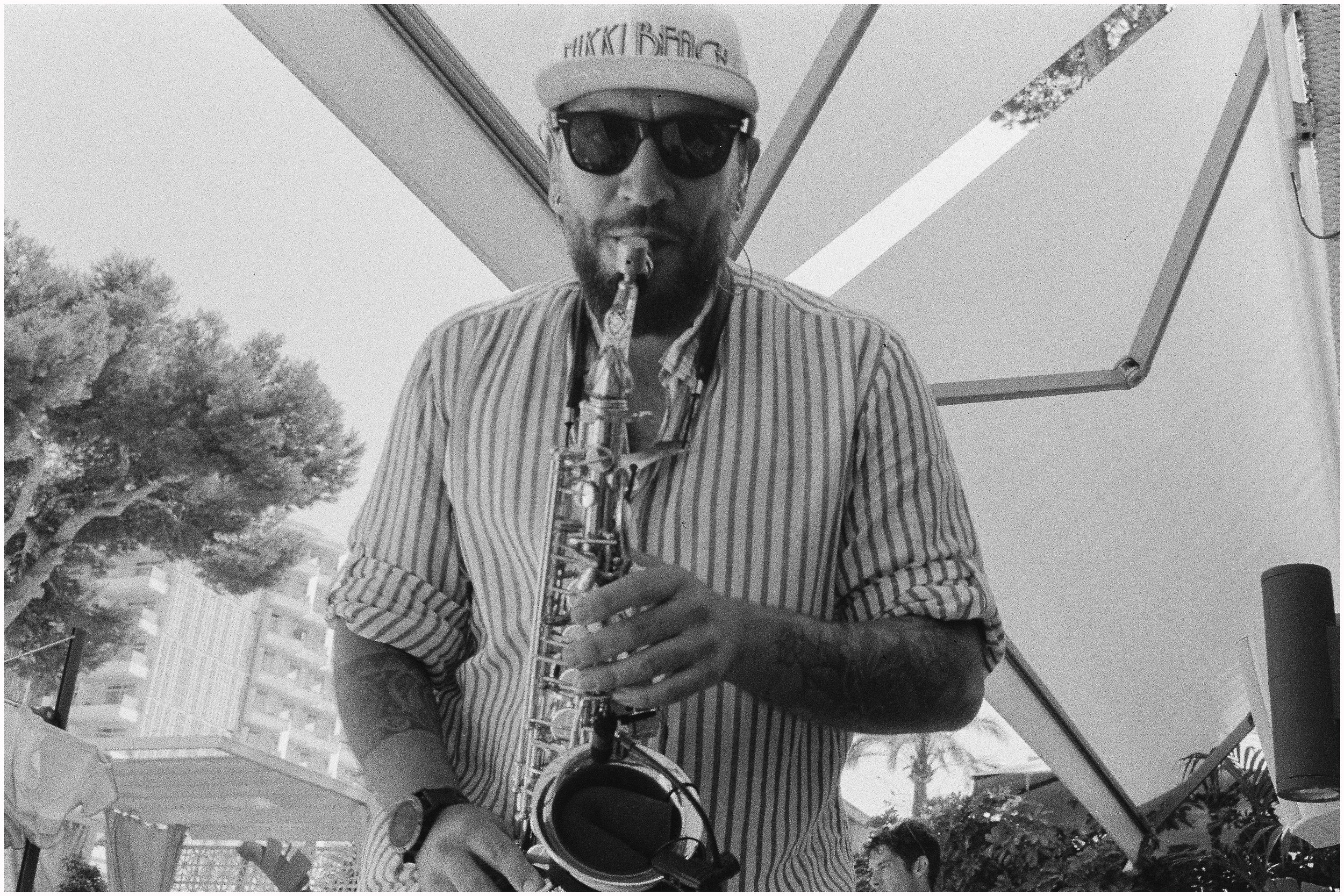 Black and white photo of a man wearing sunglasses and a cap playing a saxophone outdoors.