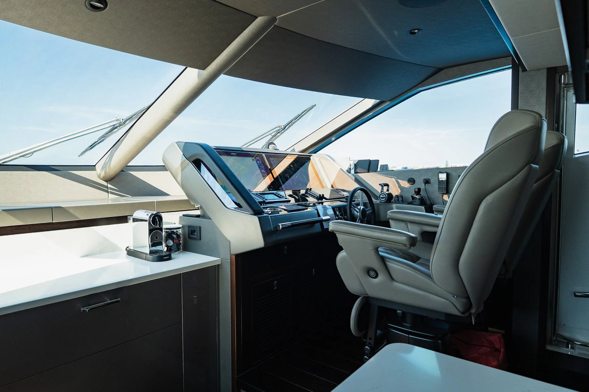 Interior view of a yacht's cockpit, featuring two beige chairs, a dashboard with navigation screens, and a coffee machine on a white countertop, with large front windows showing the sky outside.