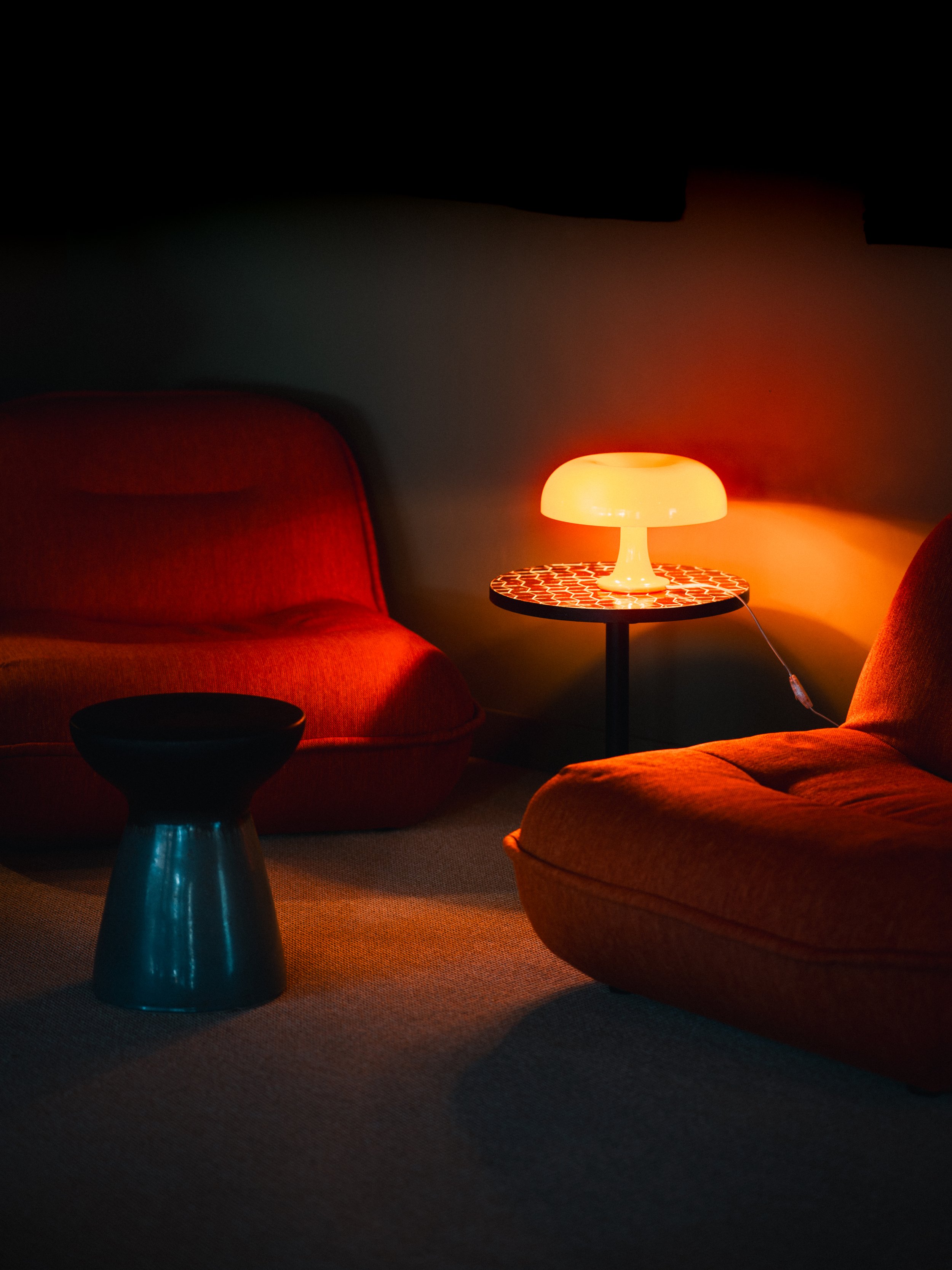 A cozy living room scene with two orange velvet chairs, a small black stool, and a glowing orange table lamp on a round side table, casting warm light.