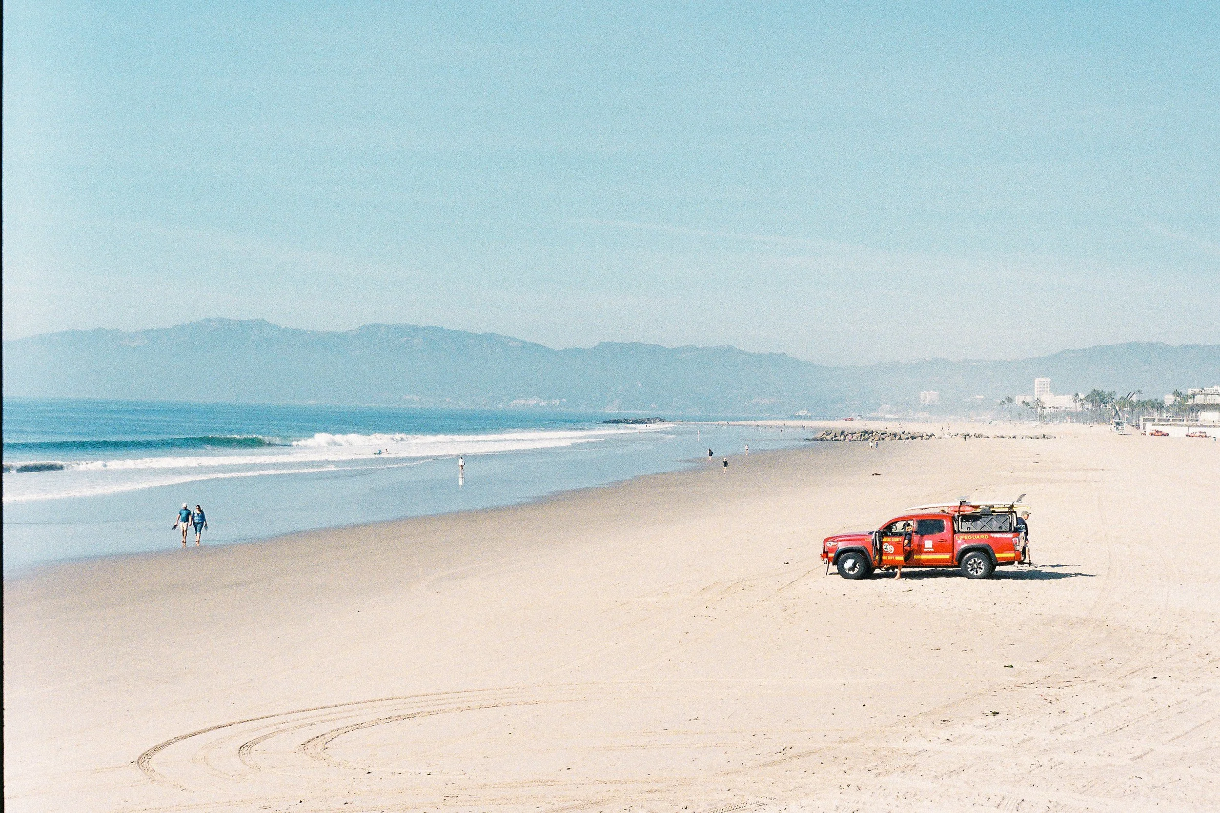 Empty sandy beach with a red lifeguard vehicle parked on the sand, a few people walking along the shoreline, and distant waves with mountains in the background under a clear blue sky.