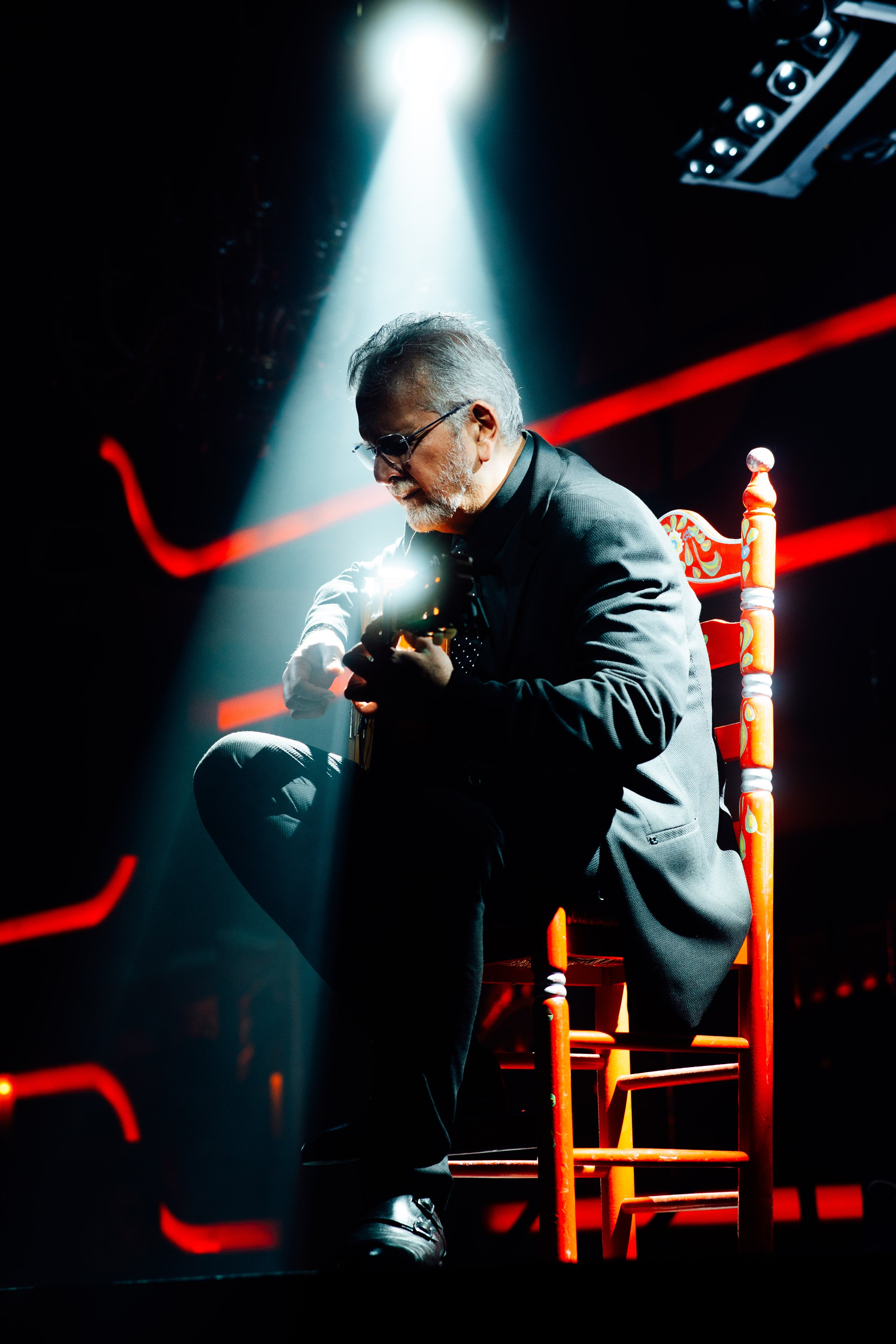 Man playing guitar on stage under spotlight with red chair and dramatic lighting.