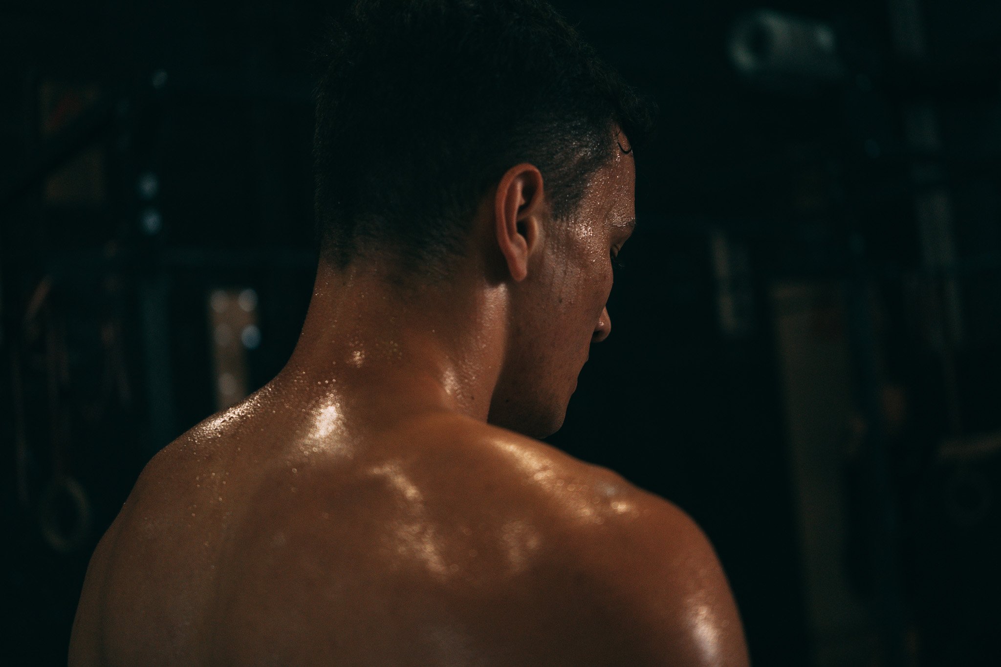 Sweaty man with short hair and muscular shoulders, looking down, in dim lighting.