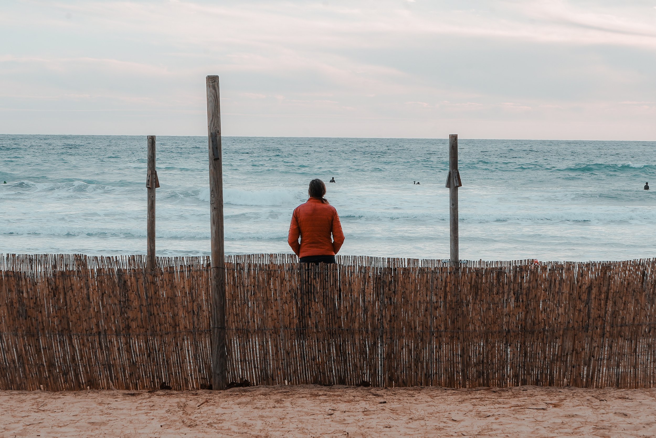 Person in red jacket standing on sandy beach behind wooden fence, looking at ocean