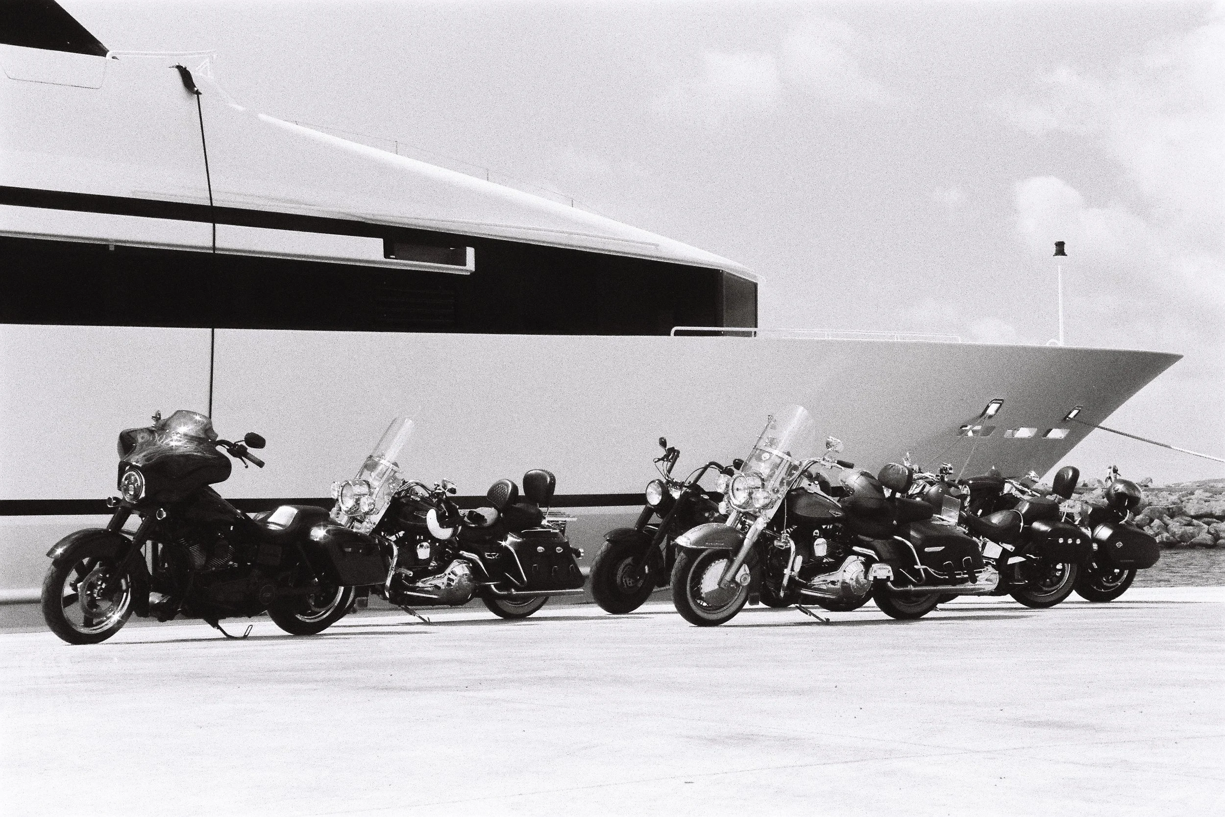 Six motorcycles parked in front of a modern building with a large, geometric overhang. The scene is in black and white and appears to be outdoors near water.