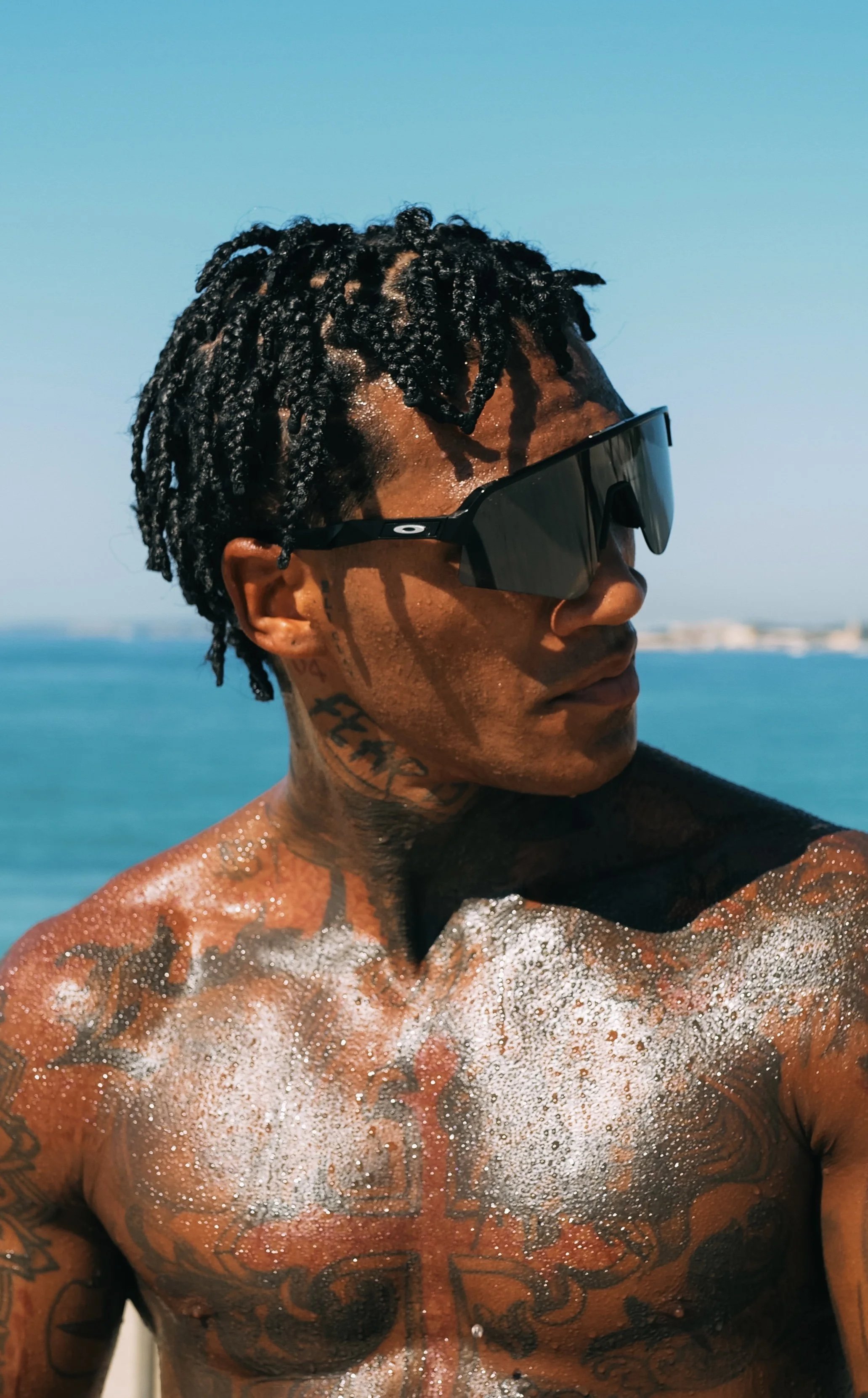 A shirtless man with tattoos and sunglasses at the beach, with water and sky in the background.
