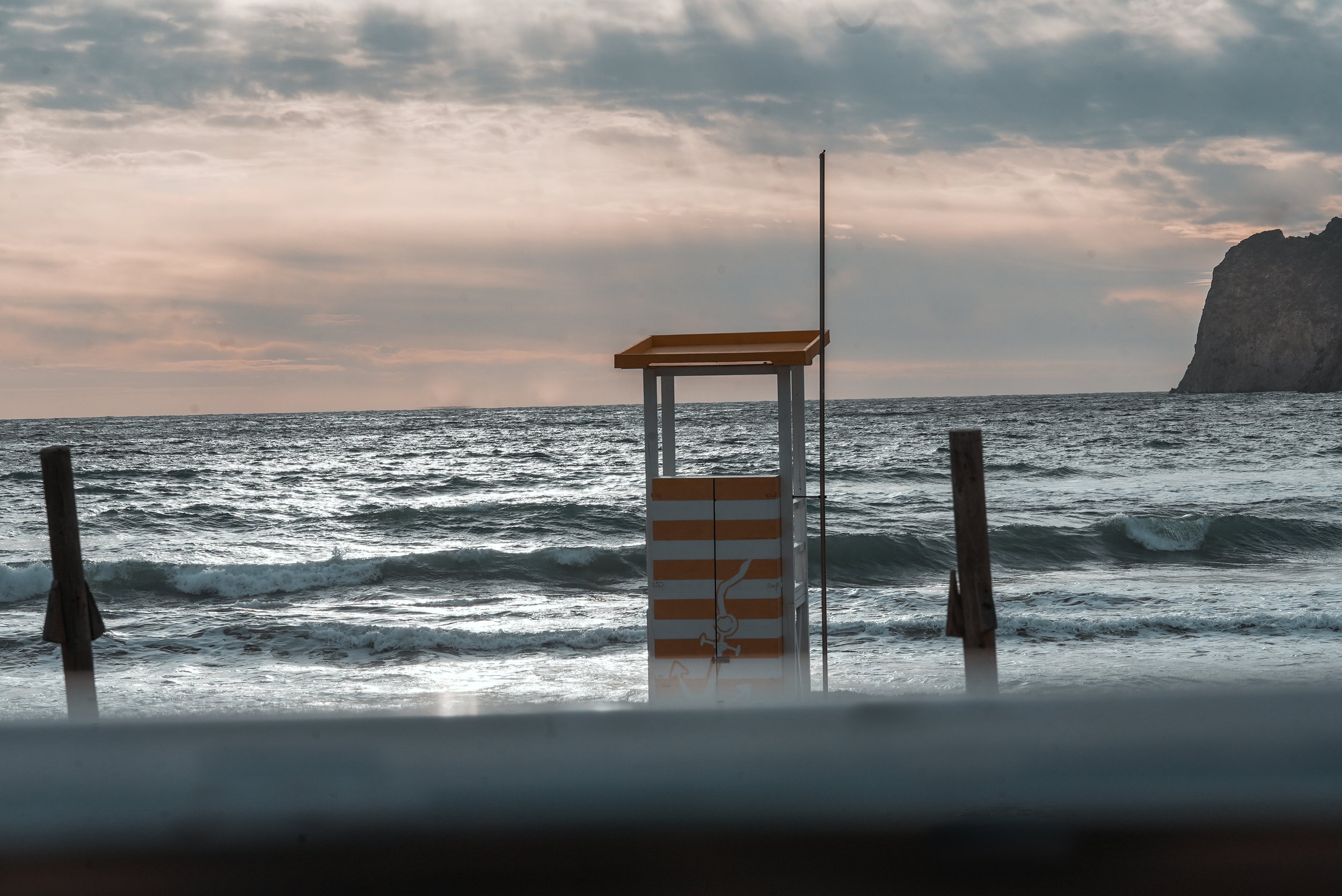 Beach scene with a striped lifeguard stand, ocean waves, and overcast sky at sunset.