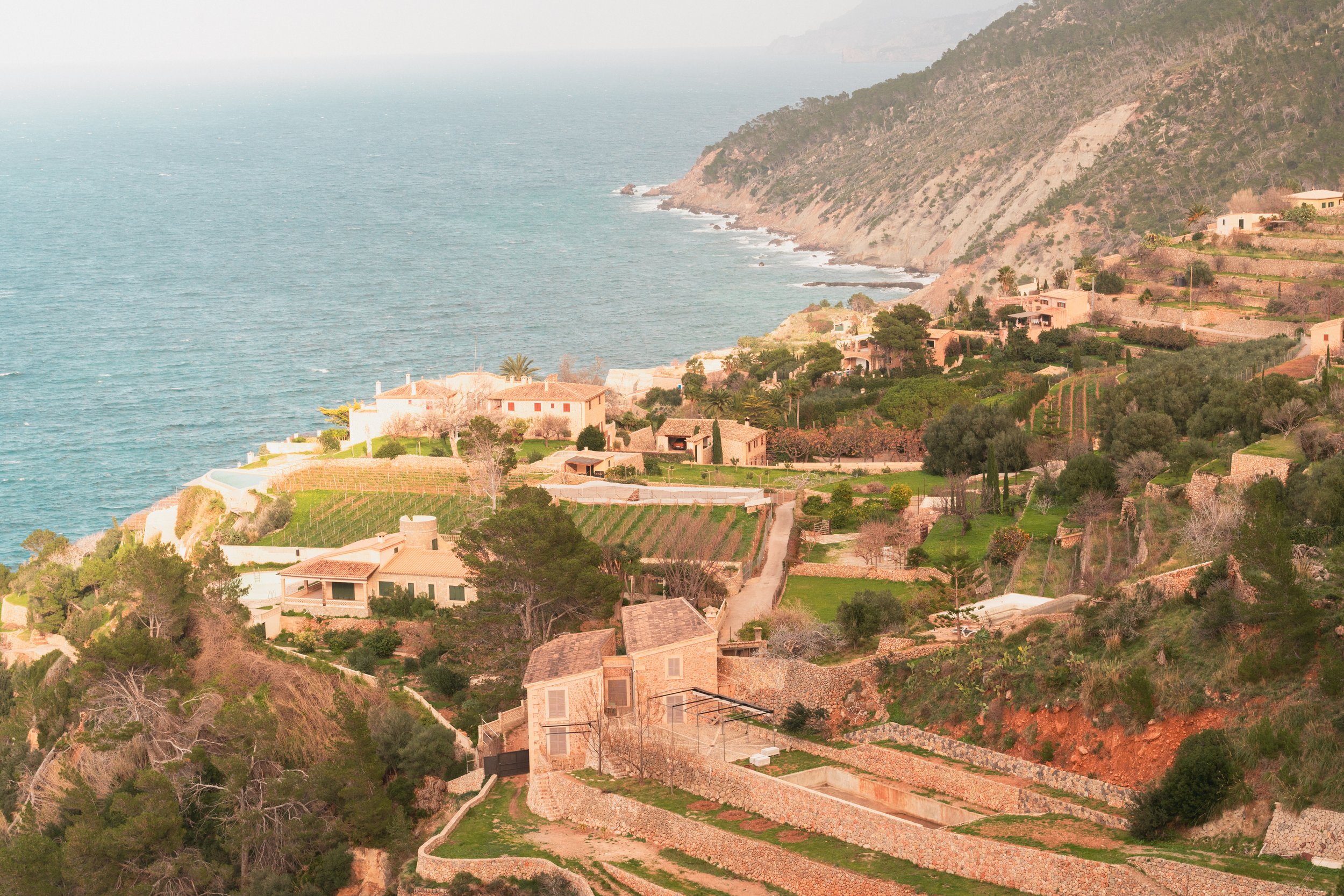 Scenic coastal view with terraced hillside, Mediterranean Sea, and buildings on the edge of cliffs at sunset or sunrise.