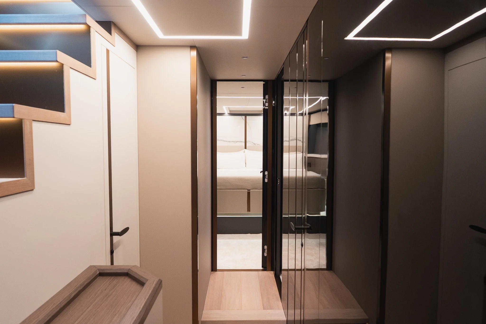Modern interior hallway leading to a bedroom with a bed and headboard visible through a glass door, features recessed ceiling lighting and wooden flooring.