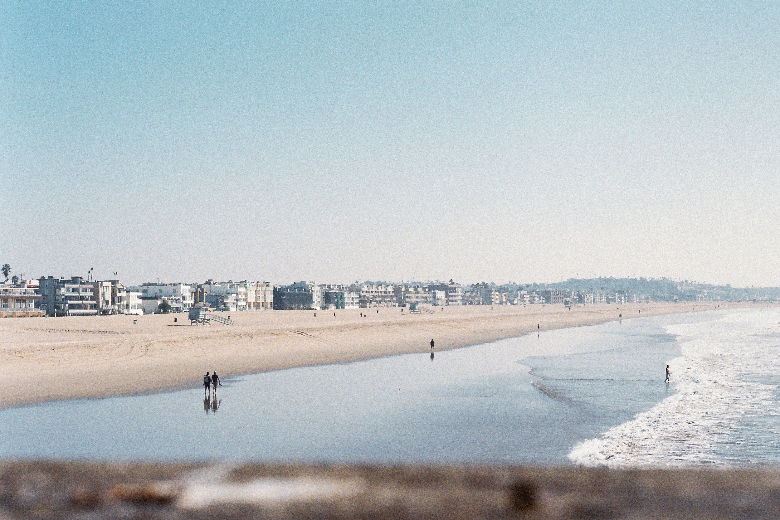 A empty beach with a few people walking along the shore, calm ocean waves, and a row of modern buildings in the background under a clear sky.