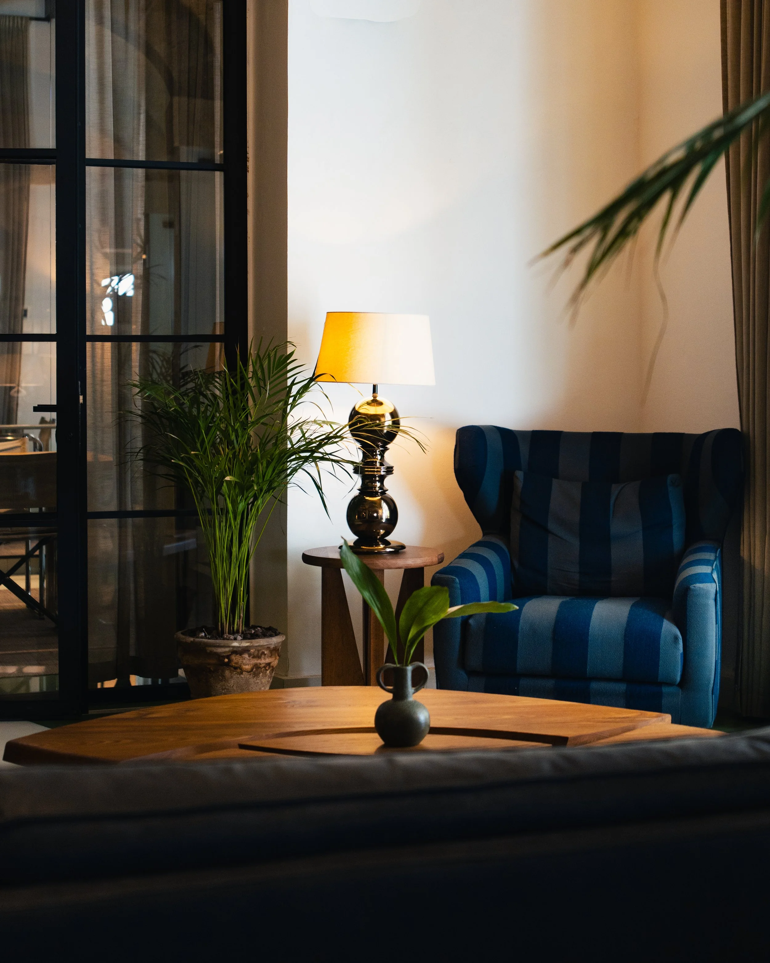 A cozy living room corner with a blue striped armchair, a small wooden side table with a black lamp, a potted plant, and a decorative vase on a wooden coffee table, with curtains and a glass door in the background.