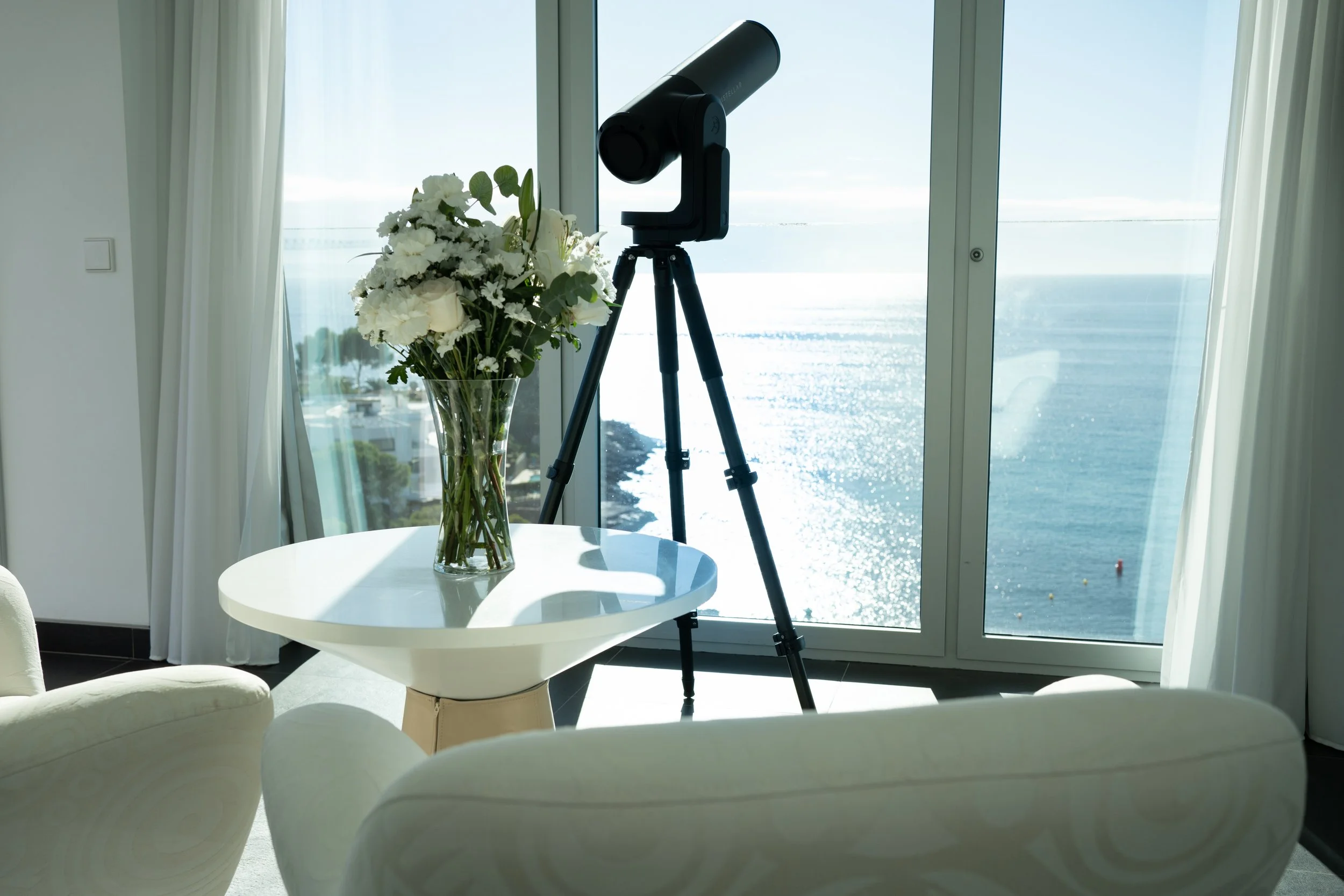 A black telescope on a tripod pointing towards a large window with a view of the ocean and sky, next to a glass vase with white flowers on a white table, in a bright room with white curtains.