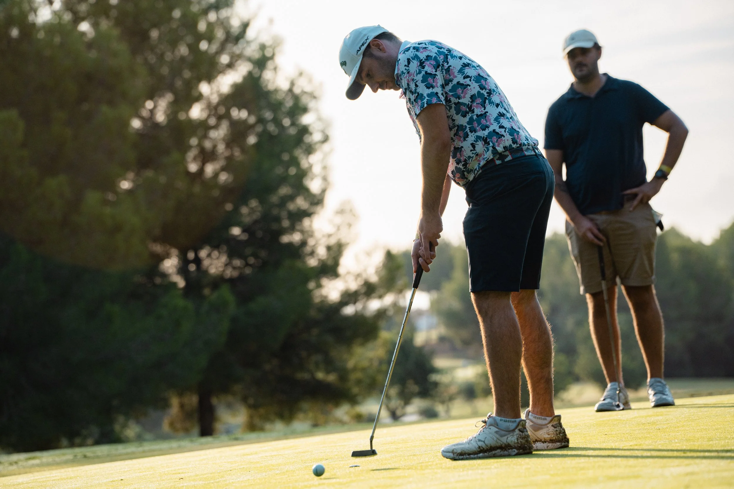 Two men playing golf on a sunny golf course, one putting and the other standing watching. The man putting is wearing a floral patterned shirt, black shorts, and a light cap. The other man is wearing a dark polo shirt, khaki shorts, and a cap, holding