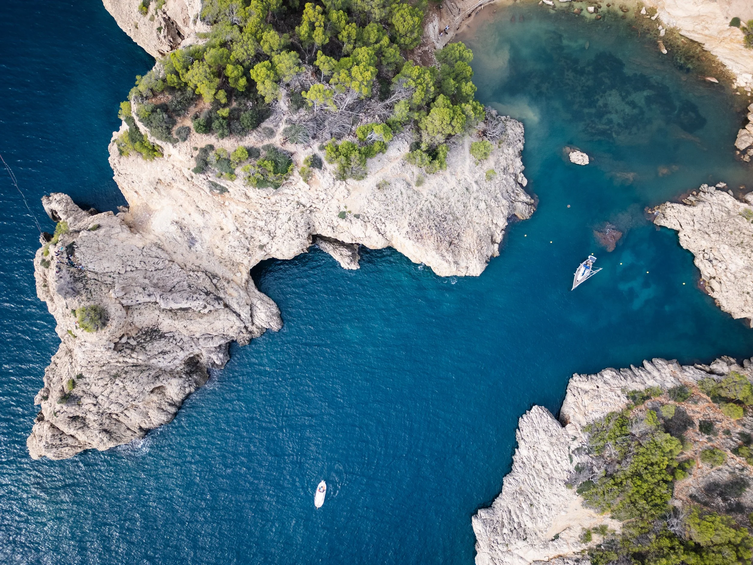 Aerial view of rocky coastline with turquoise water, trees on cliffs, and two boats in the sea.