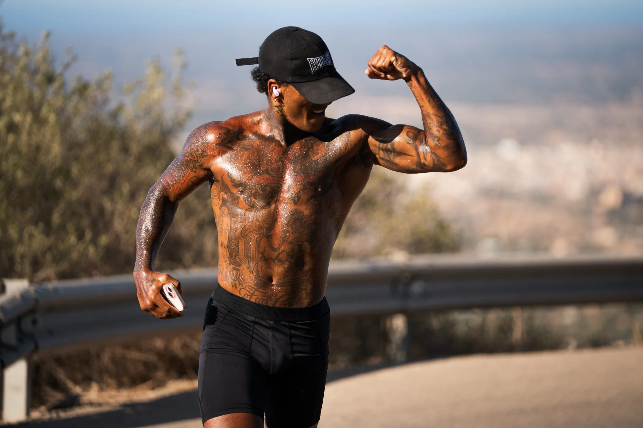 A shirtless man with tattoos in black shorts taking a selfie with a smartphone while flexing his bicep outdoors on a sunny day.