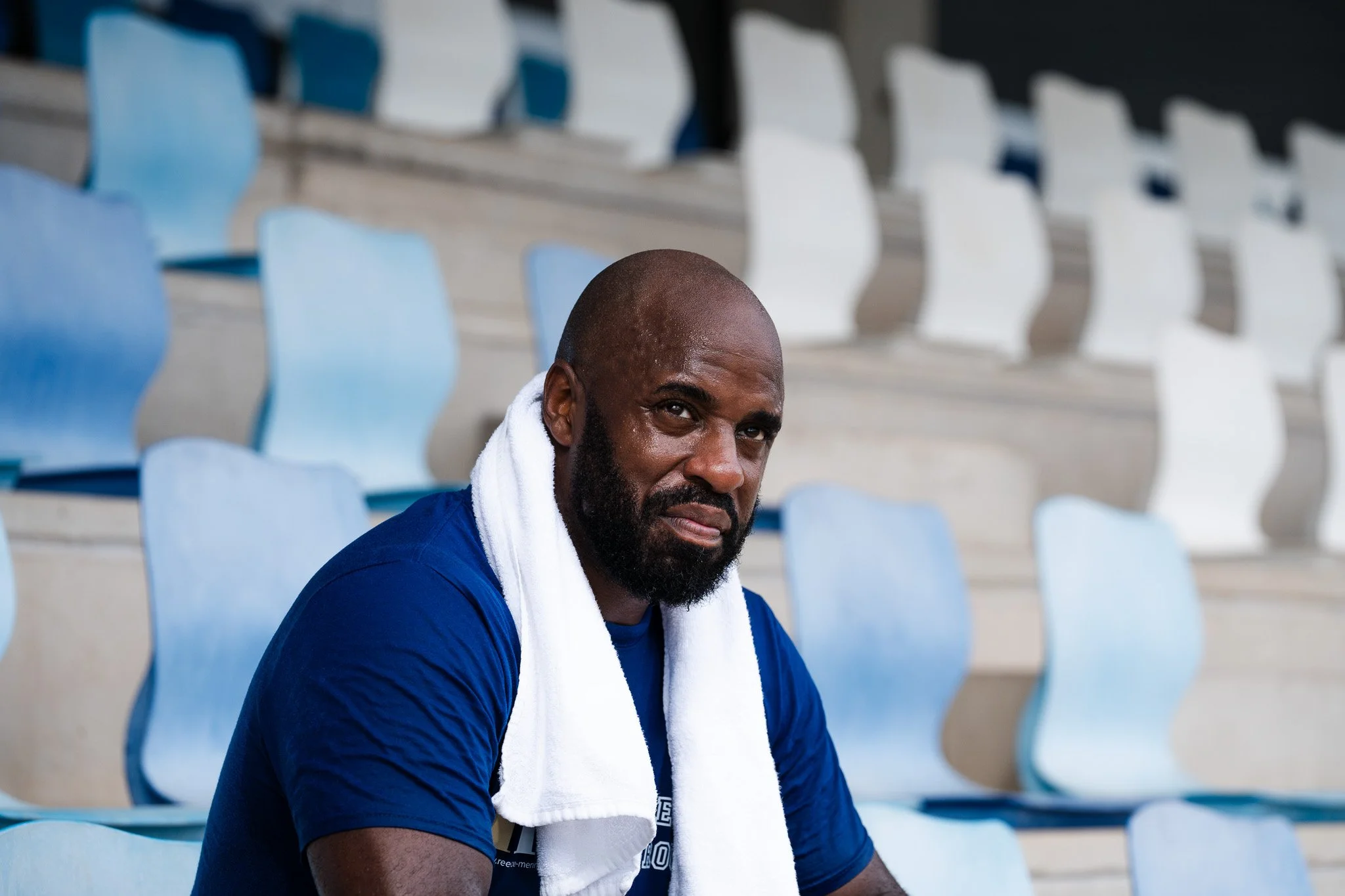 A man with a bald head and beard, wearing a blue shirt and a white towel around his neck, sitting in an empty stadium with light blue and white seats.