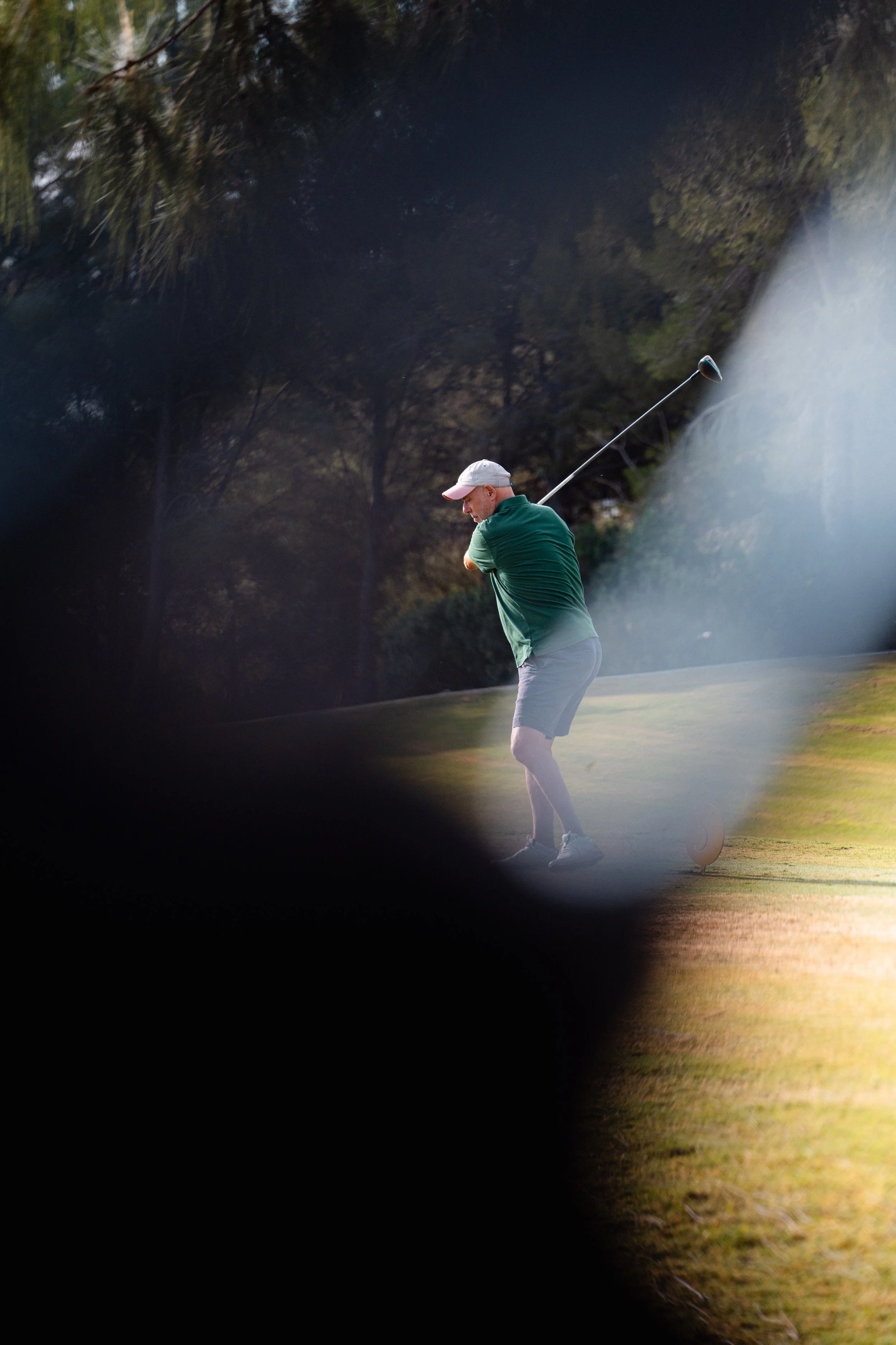 A man playing golf on a golf course, swinging his club, seen through a blurred foreground.