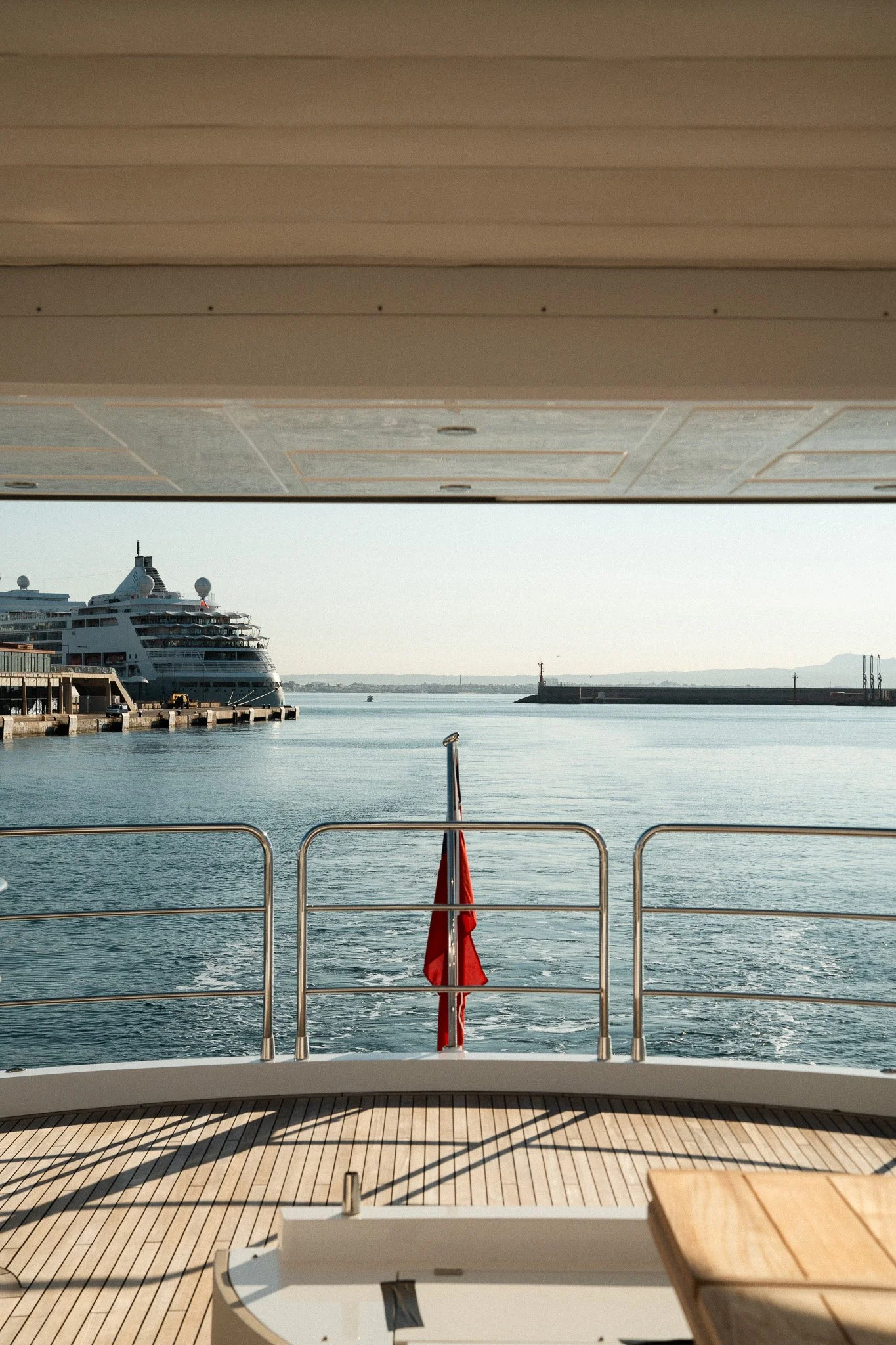 View from the deck of a yacht showing the water, a large cruise ship docked at the harbor, and distant land with a small lighthouse. A red flag is on the yacht's railing.