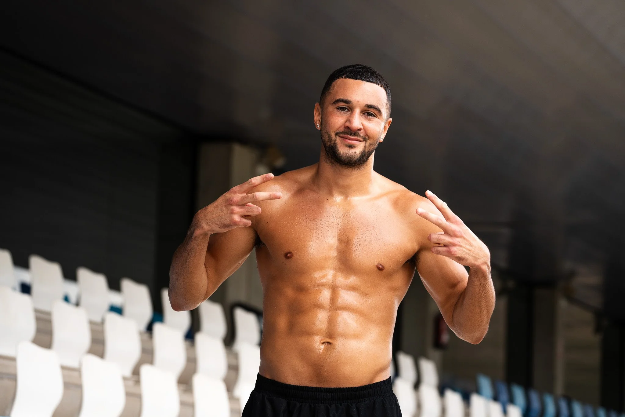 A shirtless man with short dark hair, a beard, and earrings smiling at the camera, standing in an indoor gym or sports facility with white seating in the background.