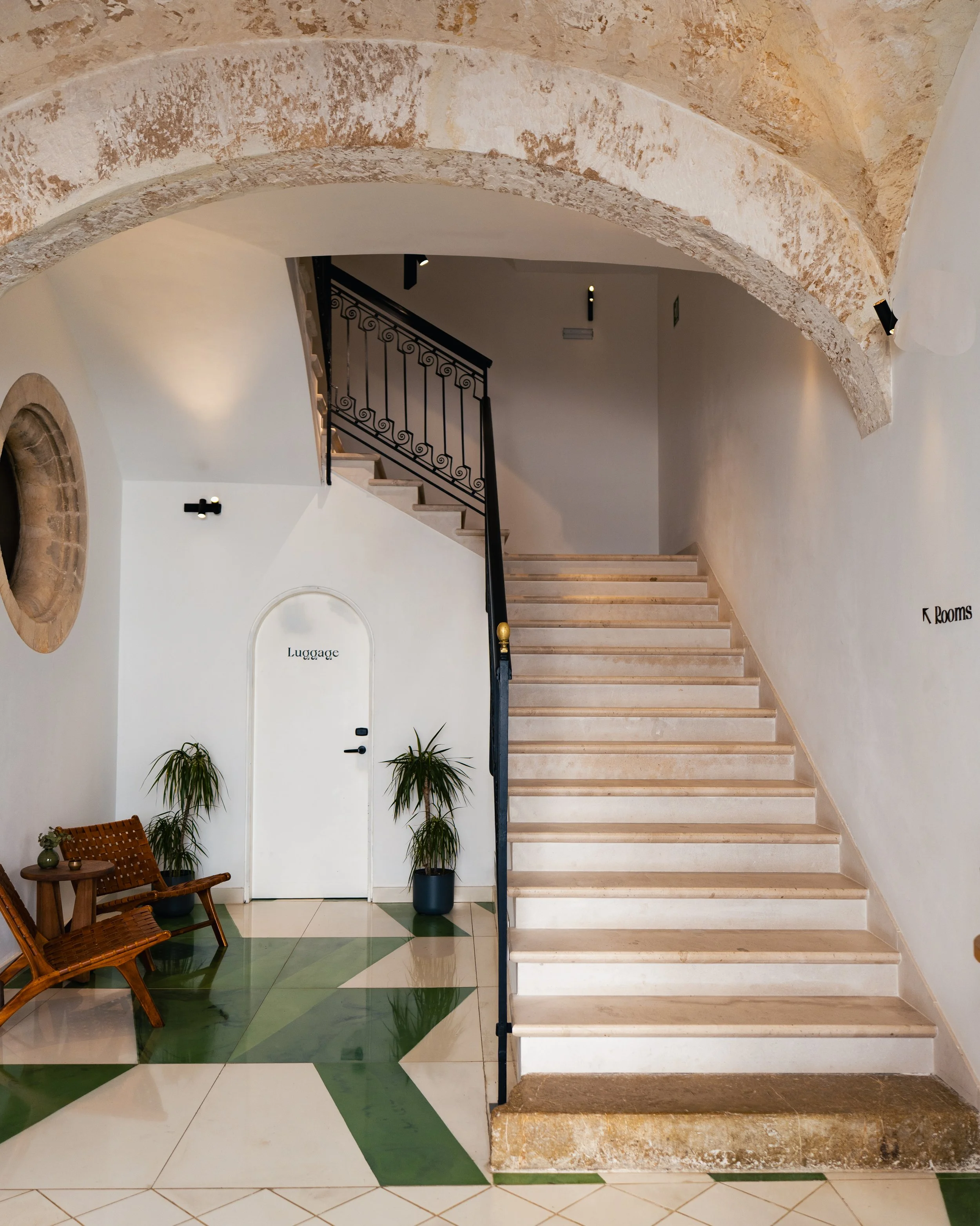 Interior of a building staircase with beige steps and a black wrought iron railing, a white wall with a small door labeled 'Luggage,' potted plants, wooden bench, and geometric green and white tile floor.