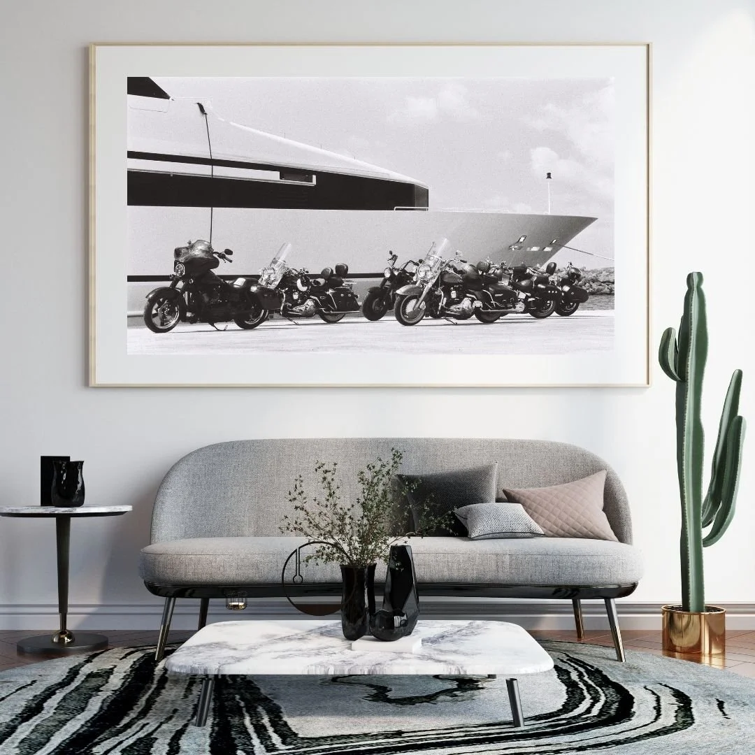 Living room with a gray sofa, a round wooden coffee table with two white cups and a book, a striped rug, and a framed picture of Harley Davidsons and a Yacht hanging on the white wall.