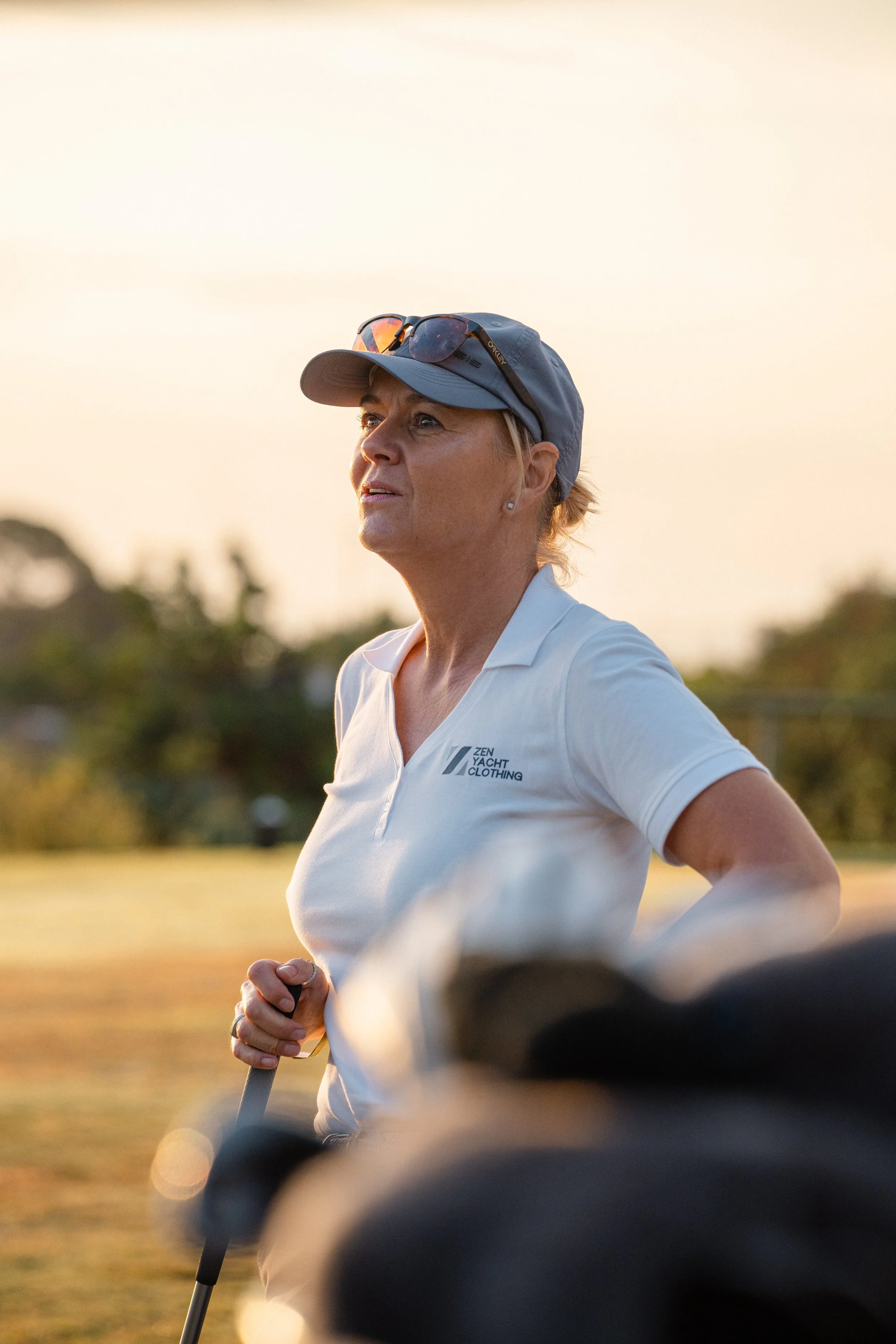 A woman in a white polo shirt and a blue cap, standing outdoors during sunset, holding a golf club, with sunglasses resting on her cap, in a golf course setting.