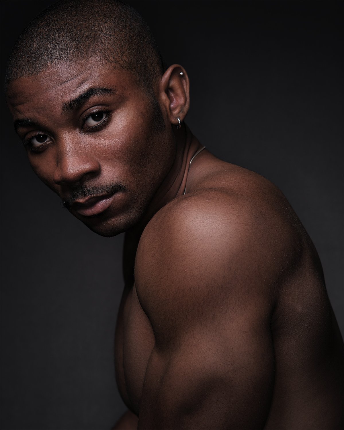 Portrait of a shirtless man with a shaved head and earring, looking at the camera, against a dark background.
