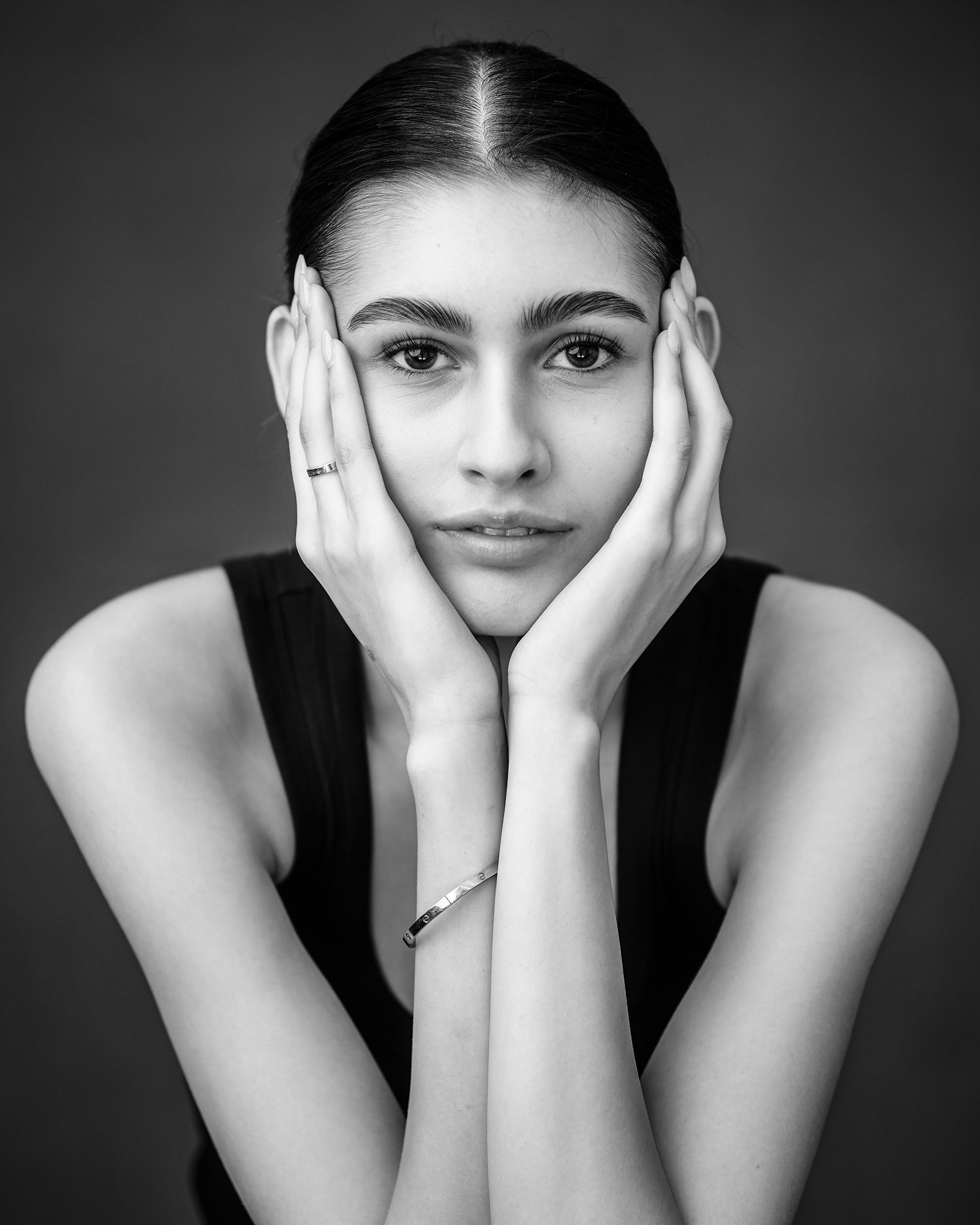 Black and white portrait of a person with hands framing their face, wearing a sleeveless top and a bracelet.