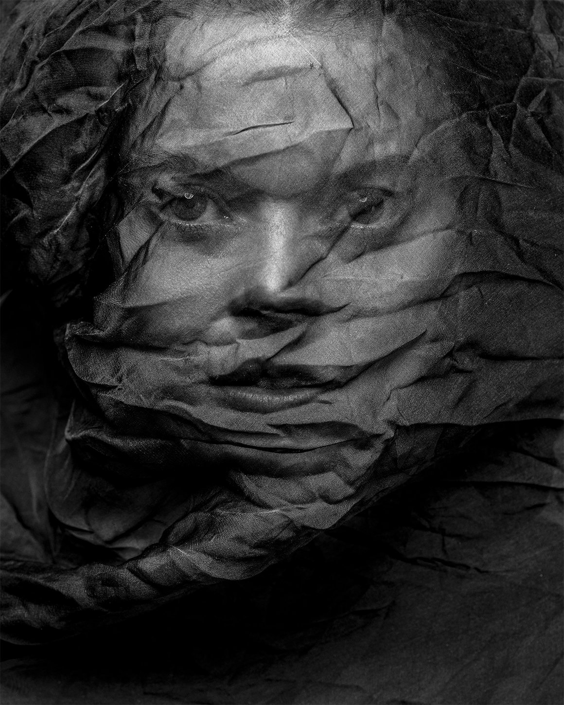 Black and white image of a person with a textured material covering their face, creating a unique visual effect with visible eyes and mouth.