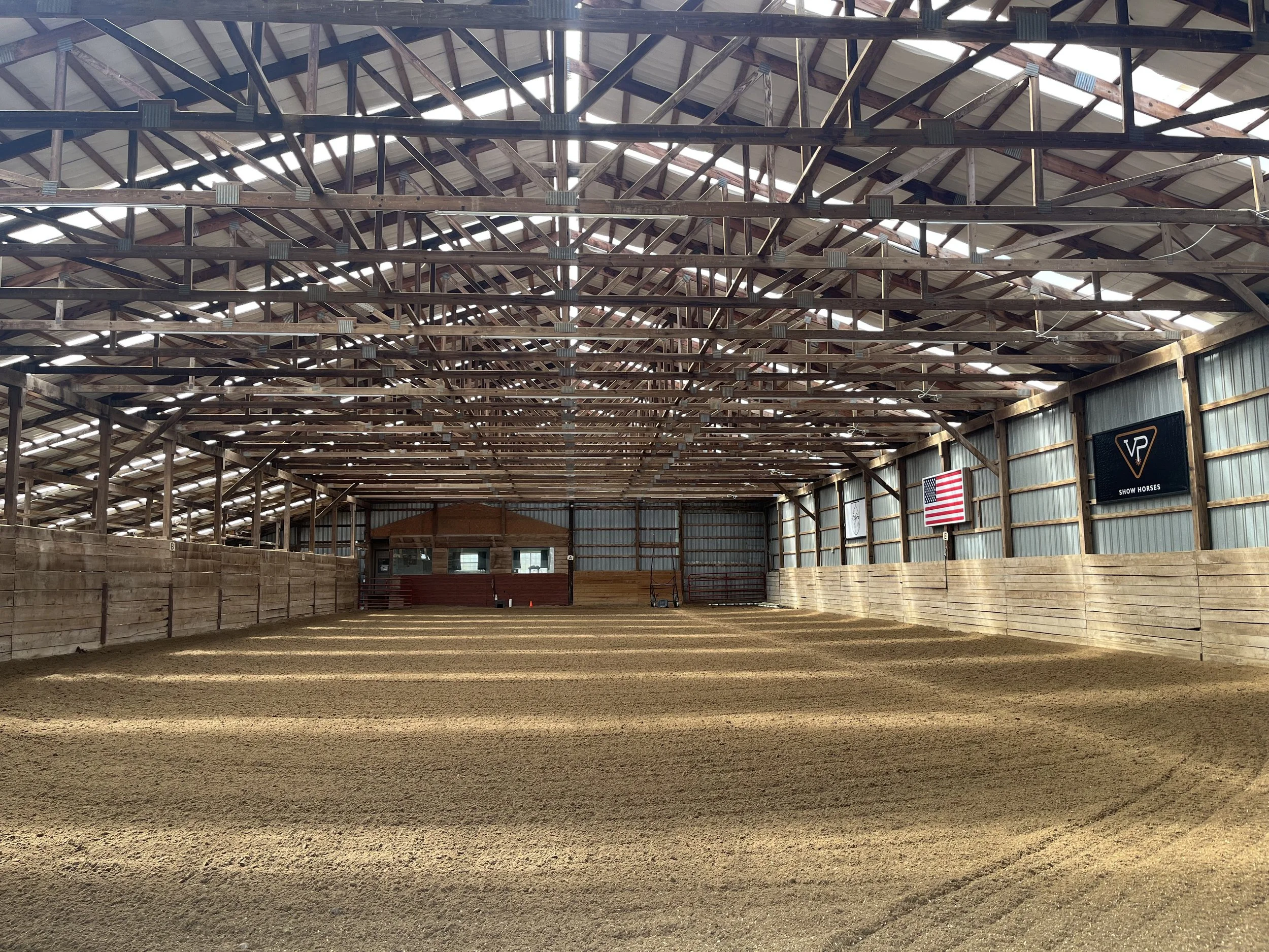 Inside an empty horse riding arena with a dirt floor, wooden railings, and a roof with exposed wooden beams and skylights.