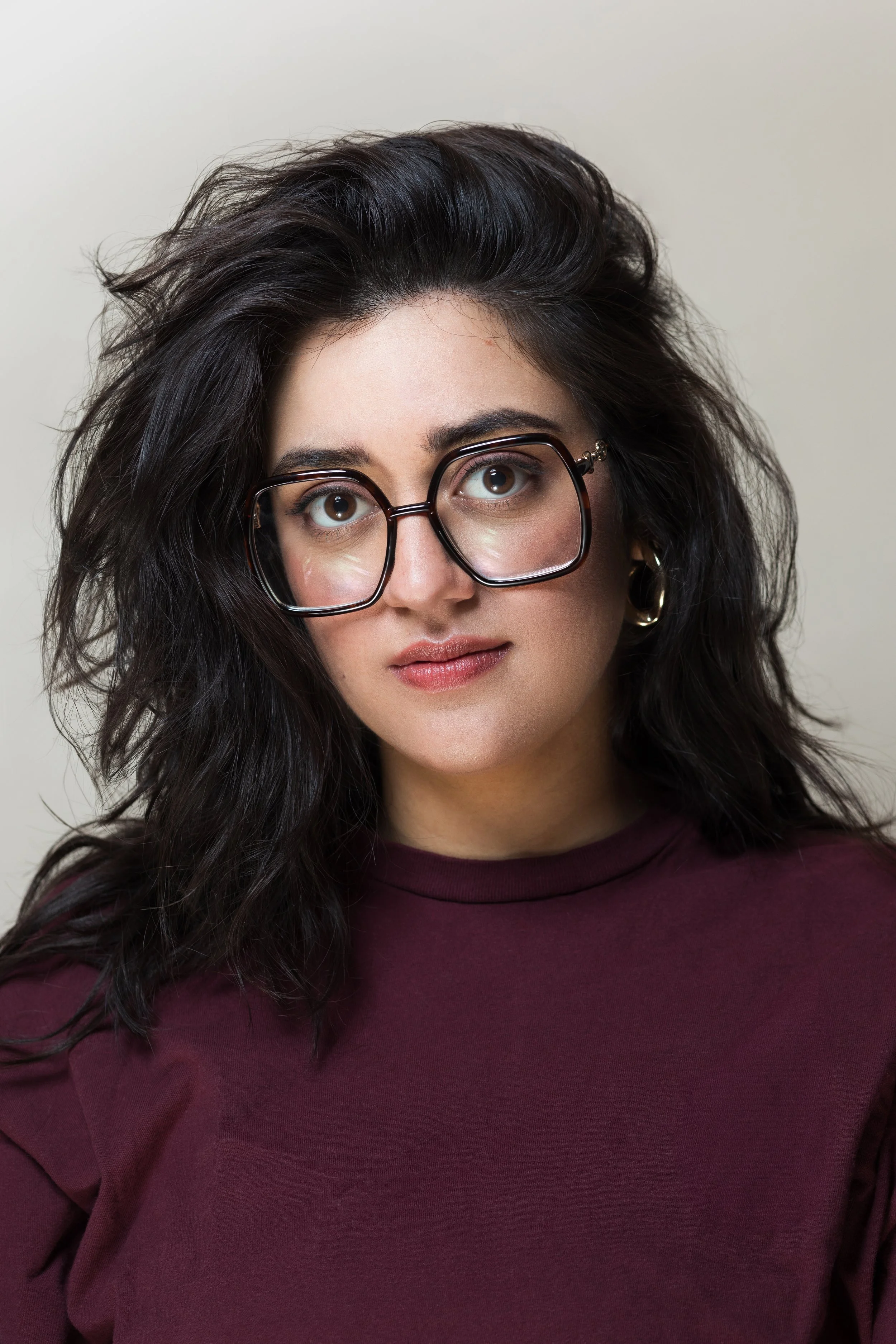A woman with curly dark hair and glasses, wearing a maroon shirt, looking at the camera with a neutral expression.