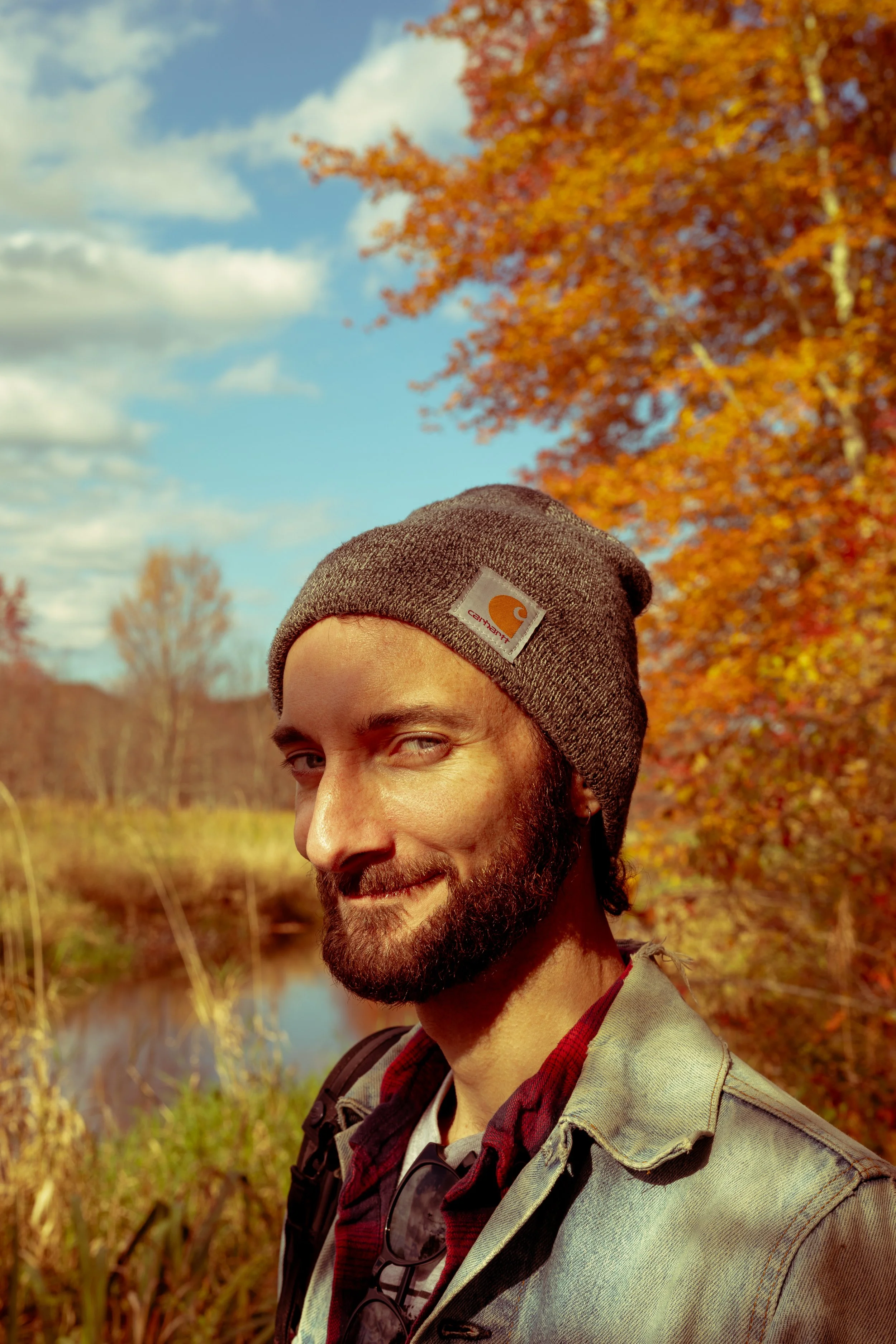 A man with a beard, wearing a gray beanie and a denim jacket, smiling outdoors during autumn in a wooded area with colorful fall foliage and a small river in the background.