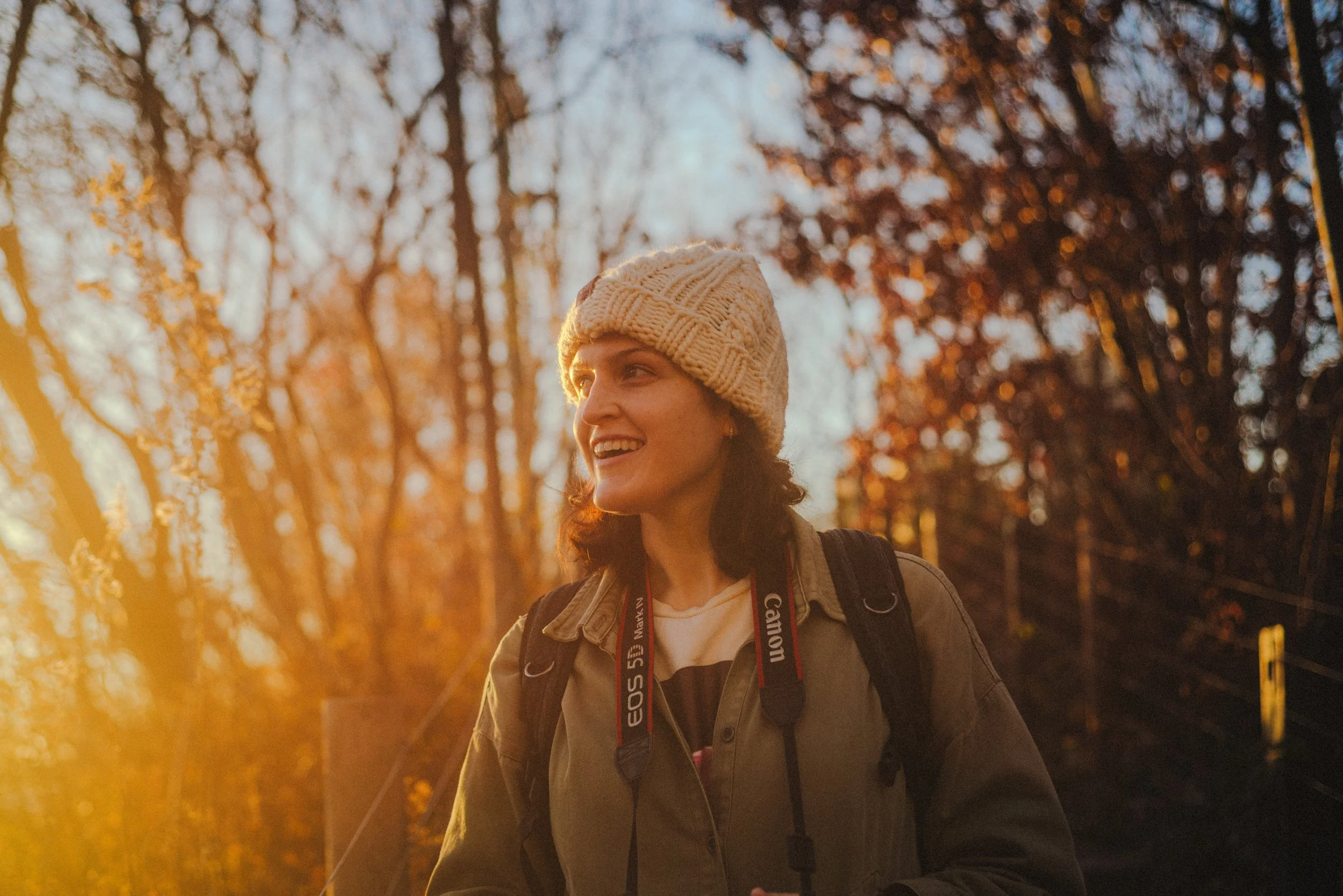 A woman smiling outdoors during sunset in a wooded area, wearing a beige knit hat, a khaki jacket, and carrying a camera around her neck and a backpack.