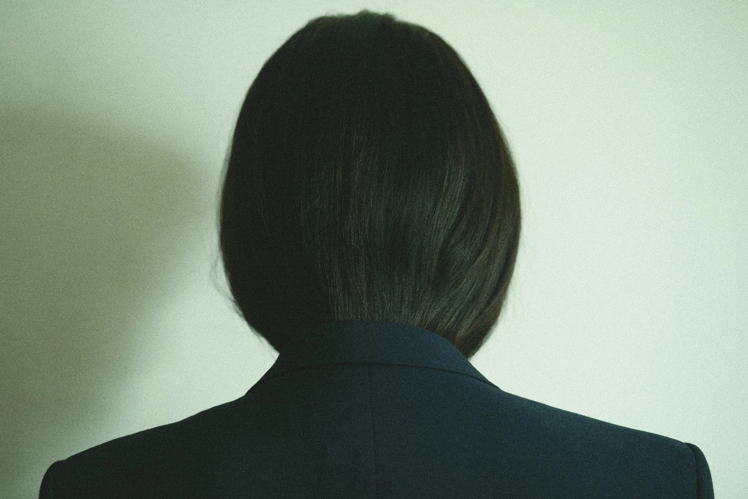 Back of a person's head with dark brown hair and a black blazer, against a plain light-colored wall.