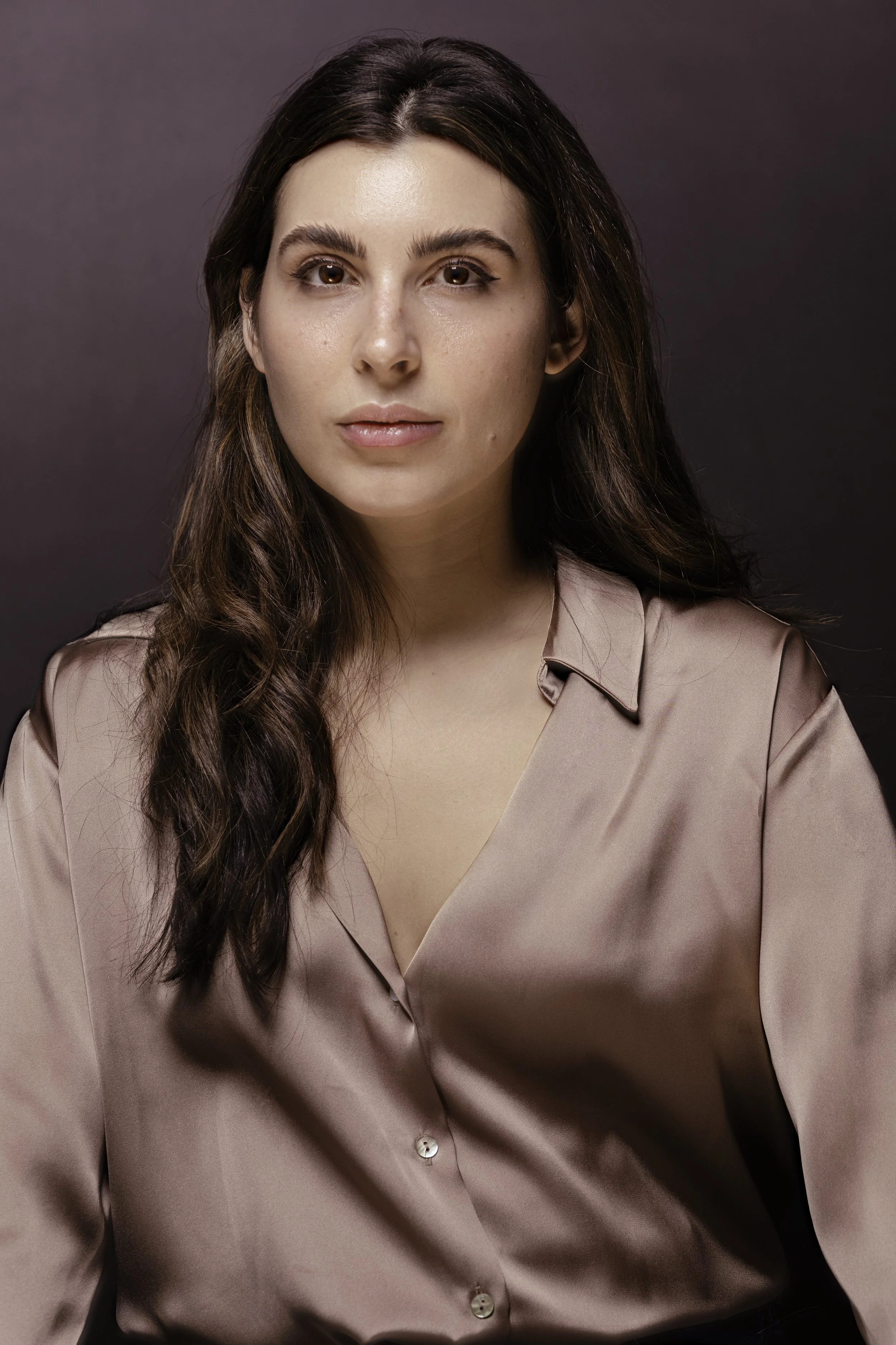 A woman with long, wavy brown hair and light skin, dressed in a silky beige blouse, sitting against a dark background.