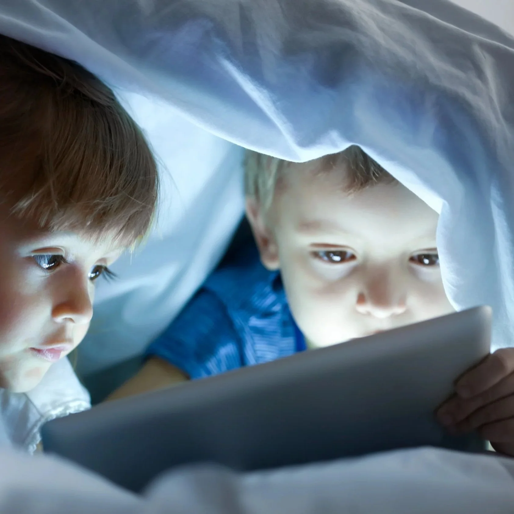 Two young children under a blanket playing on a tablet.