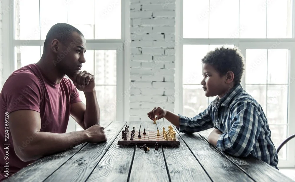 Father and 10 year old son playing chess on wooden table in front of large windows.