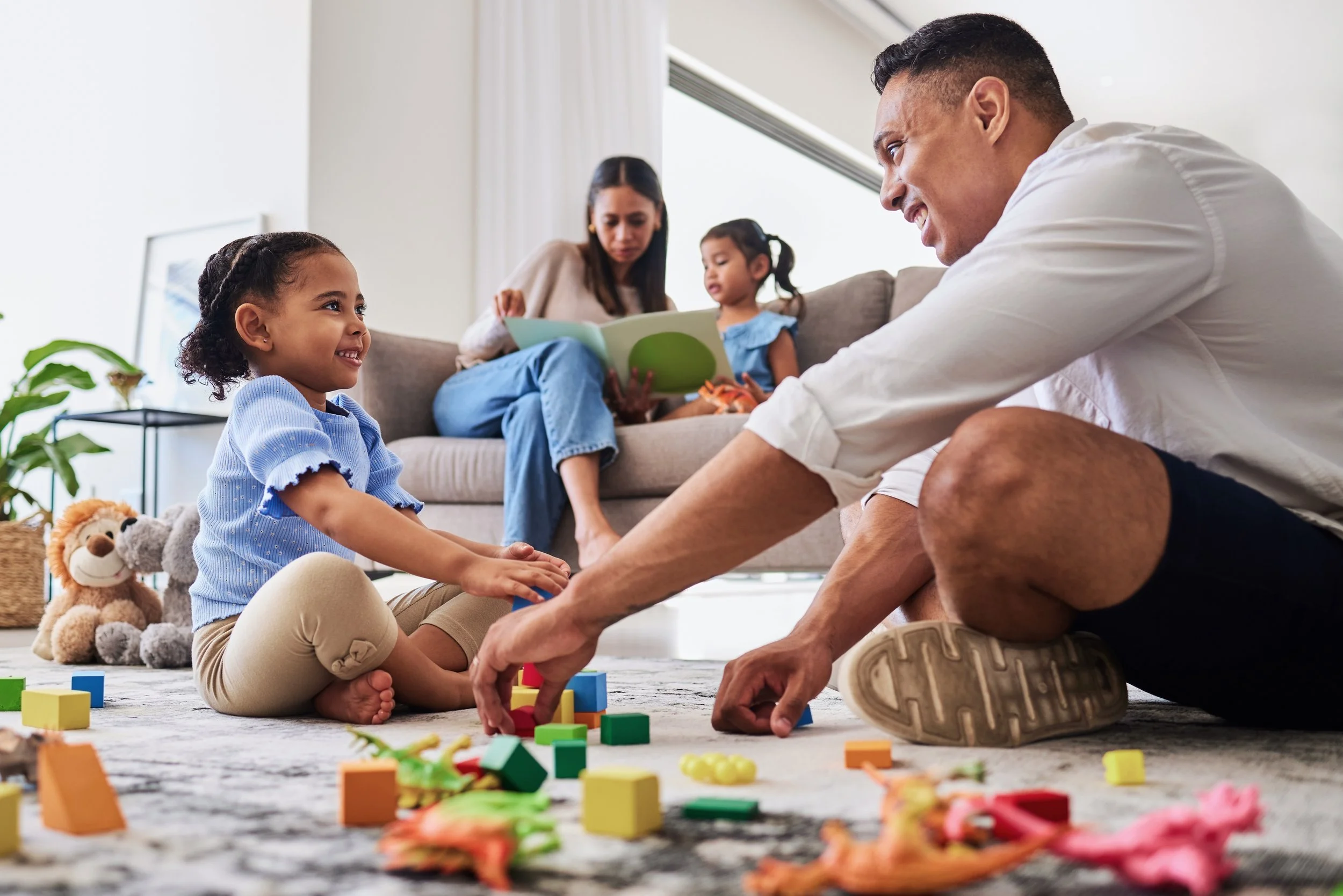 Father playing with blocks with his son on the floor while mother and daughter read on the couch.